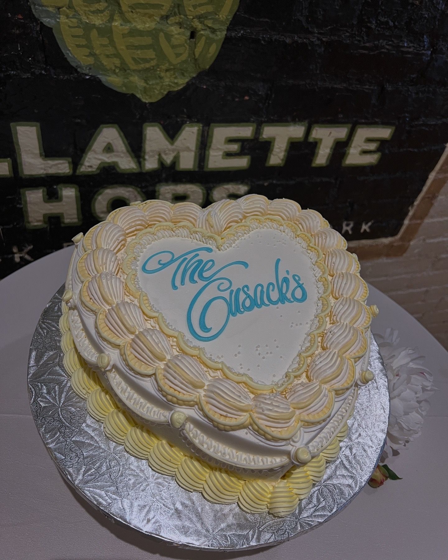 A heart-shaped white and yellow frosted cake decorated with 'The Classics' text sits on a silver cake board.