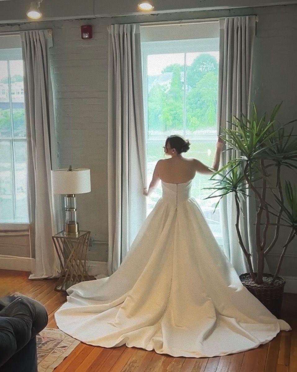 Elegant strapless white wedding gown with a dramatic train shown from behind in a sunlit room with tall windows.