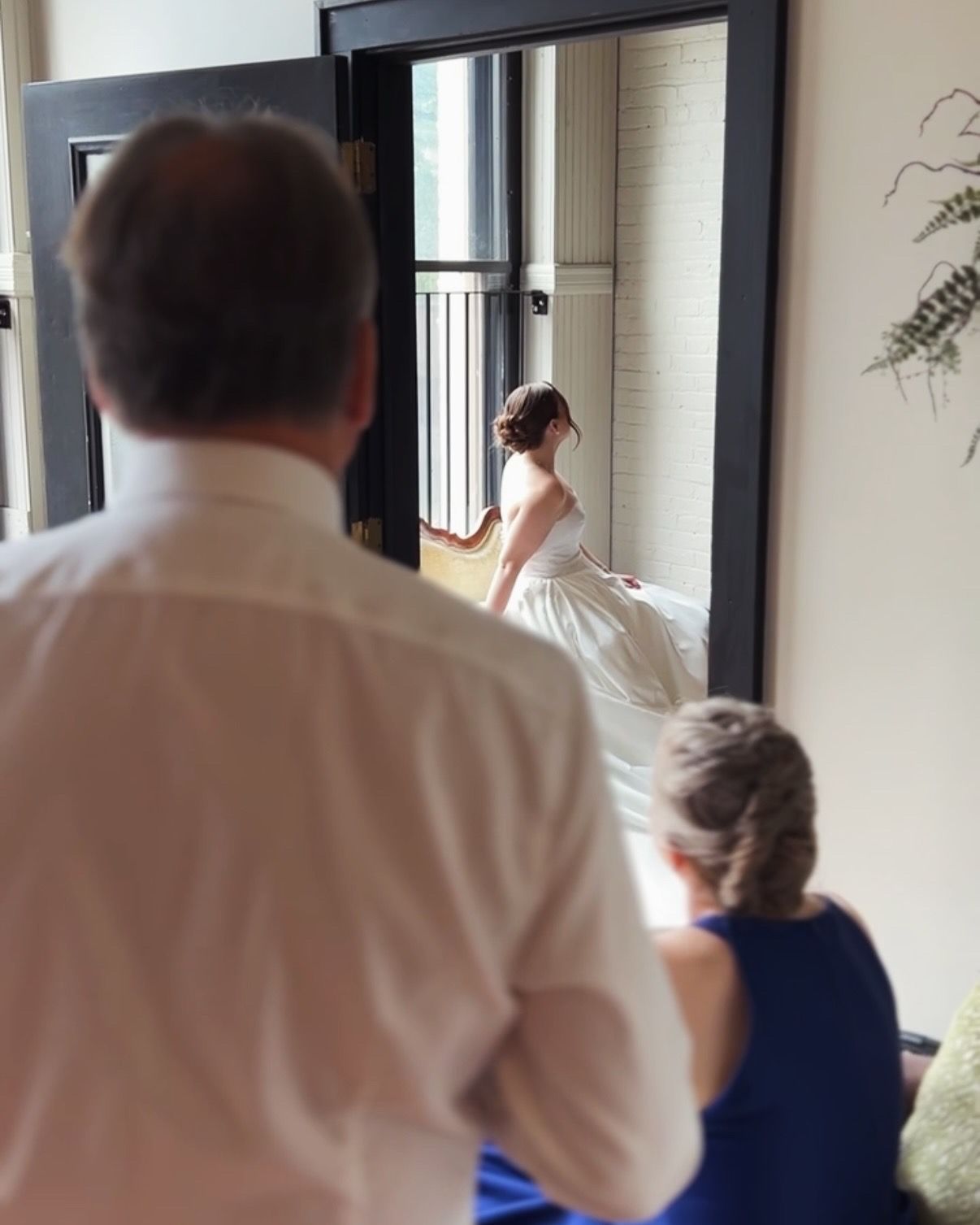 View through doorway of bride in white gown looking out large window in bright sunlit room.