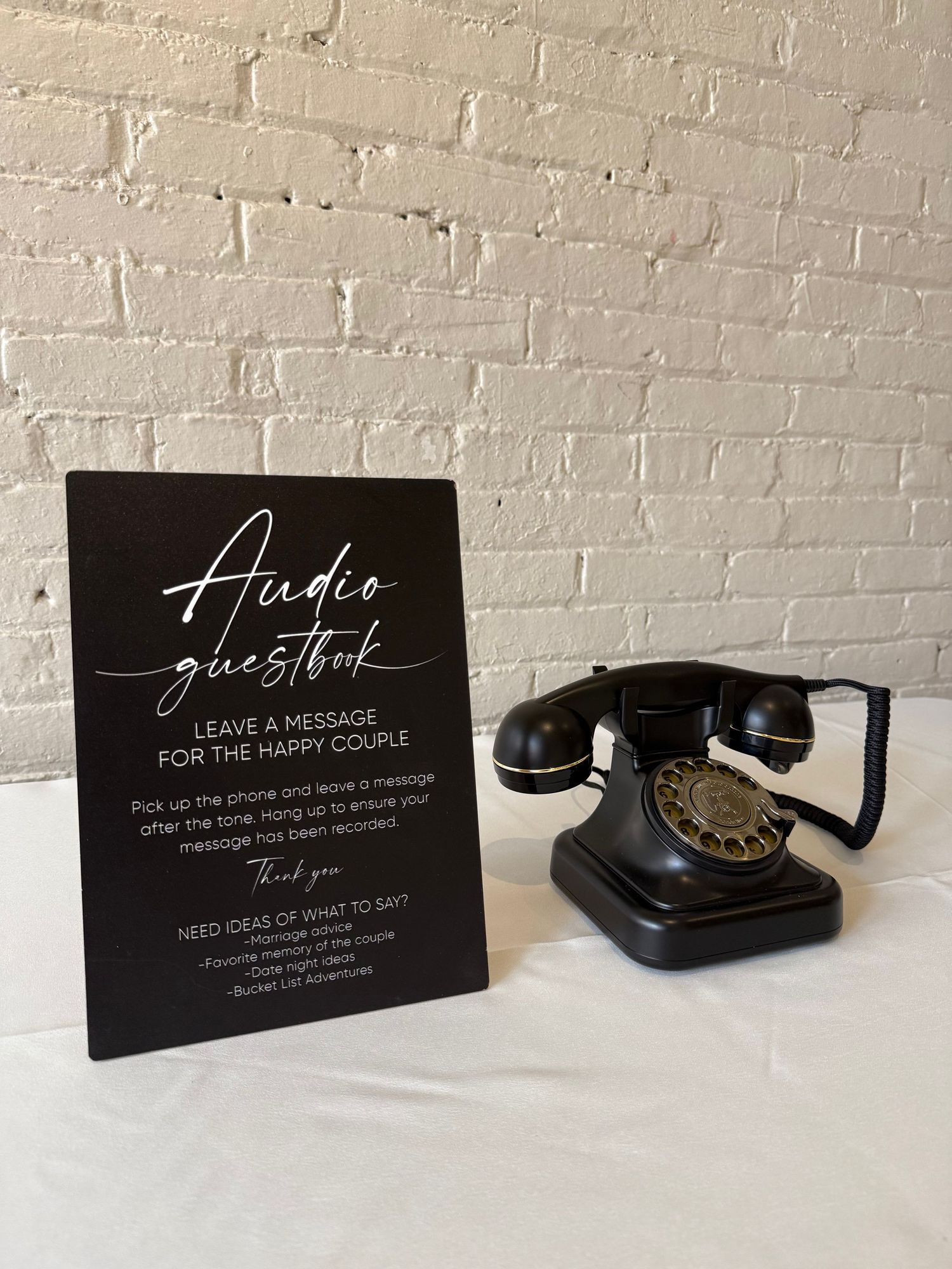 A vintage black rotary telephone sits beside black acrylic guest book signs with white lettering on a white surface.