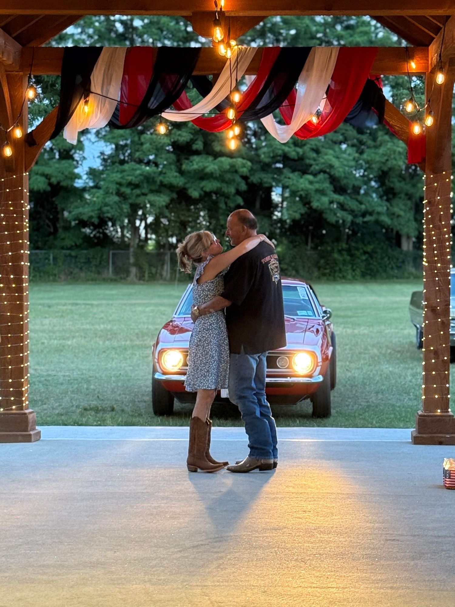A couple sharing a dance at dusk under a rustic pavilion with string lights and red drapes, while a car's headlights glow behind them.