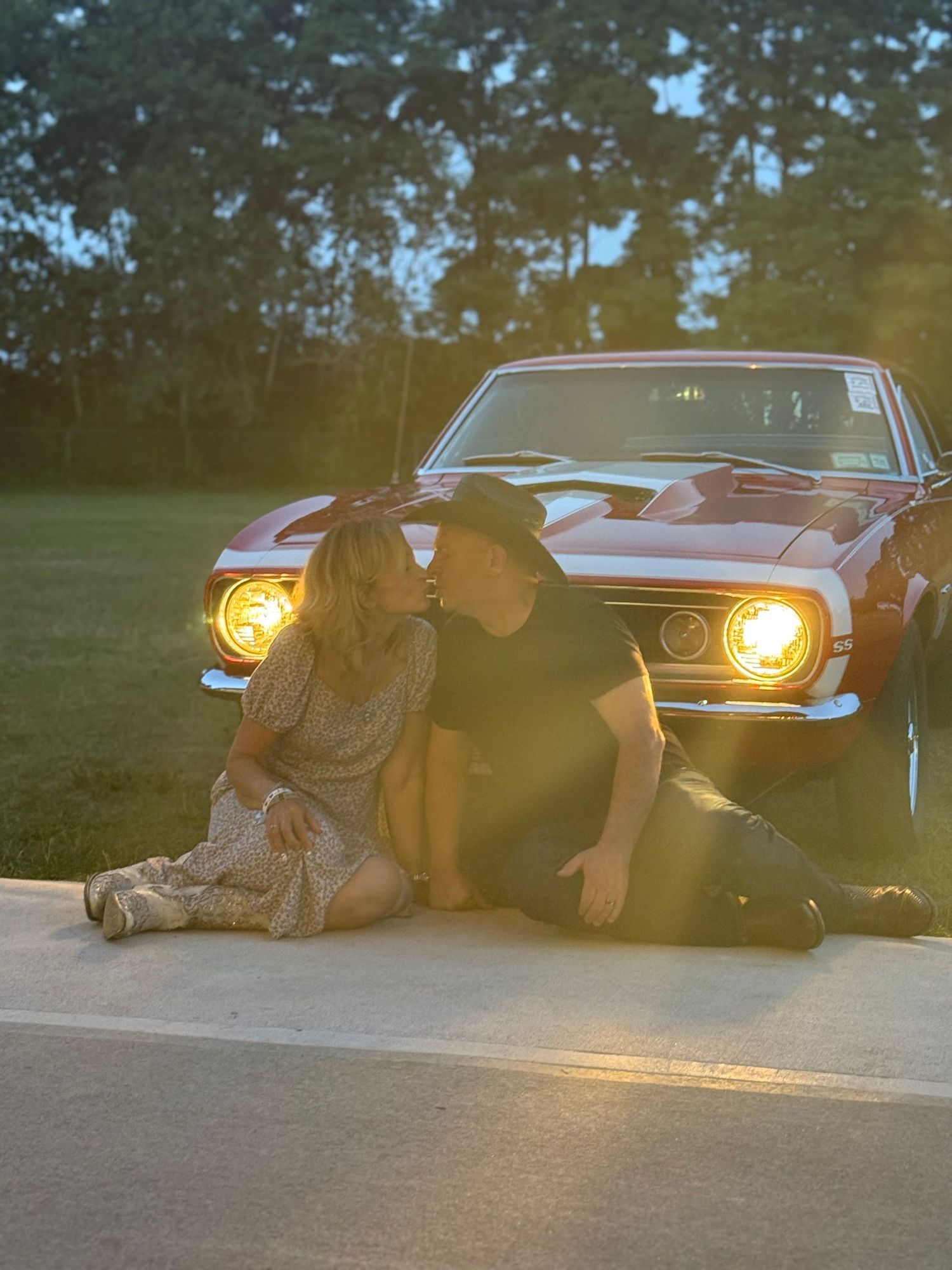 A vintage muscle car with glowing headlights parked at dusk on a summer evening.