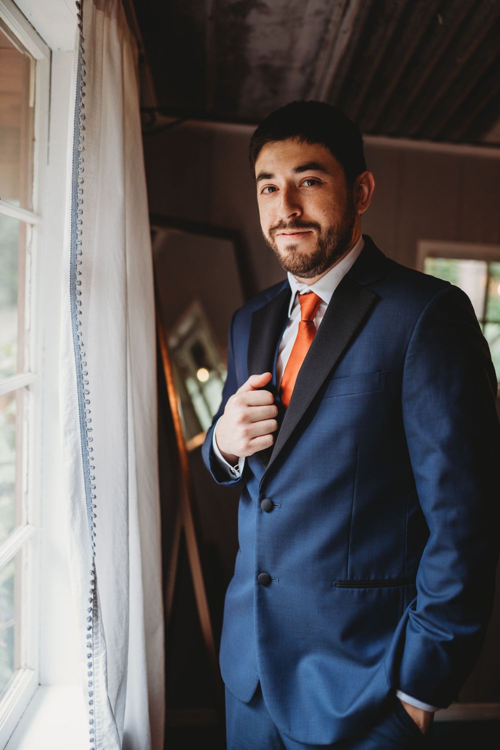 Formal portrait of gentleman in navy suit with orange tie standing by window in natural light.