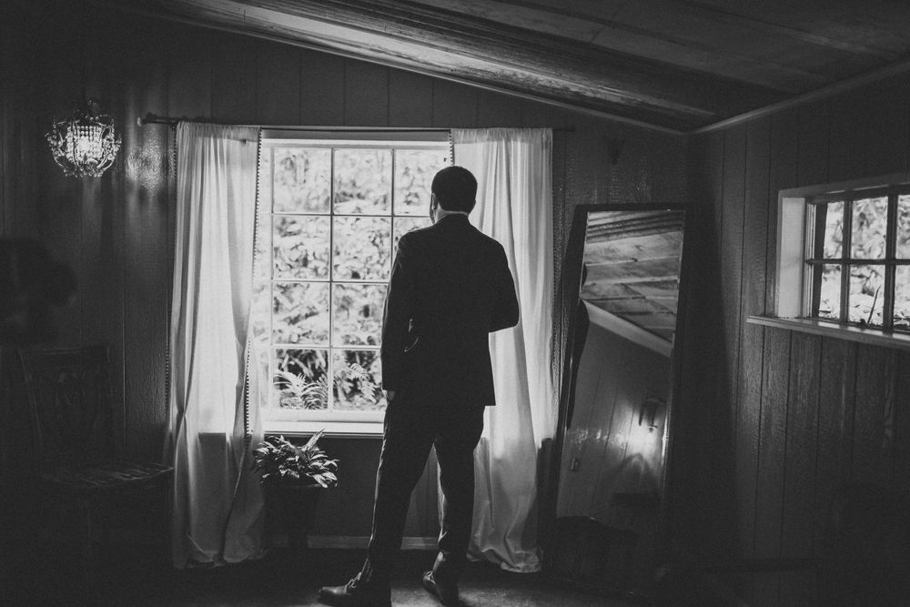 Silhouetted figure stands contemplatively by window in moody black and white photograph.