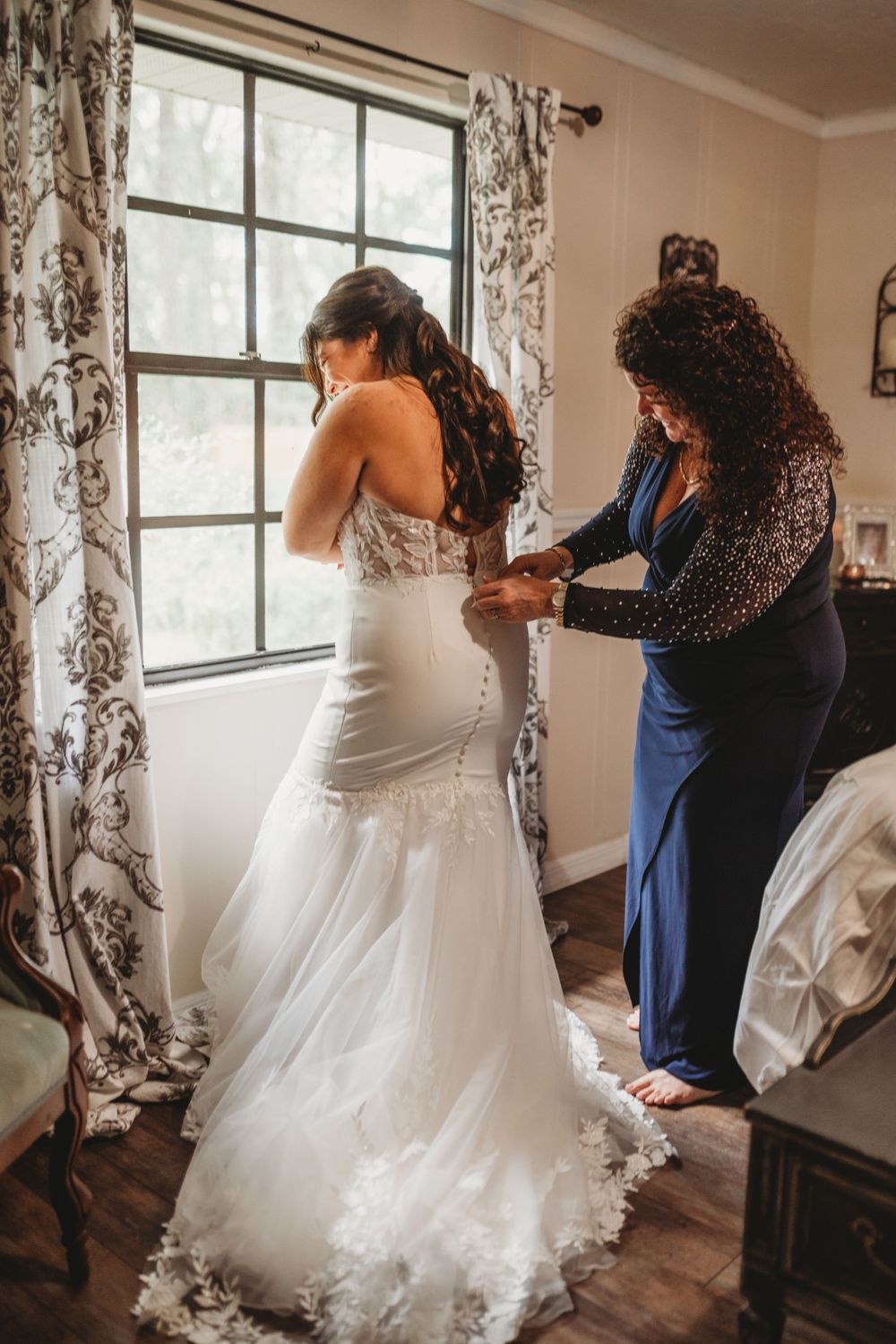 Final wedding dress adjustments being made near sunlit window in preparation room.