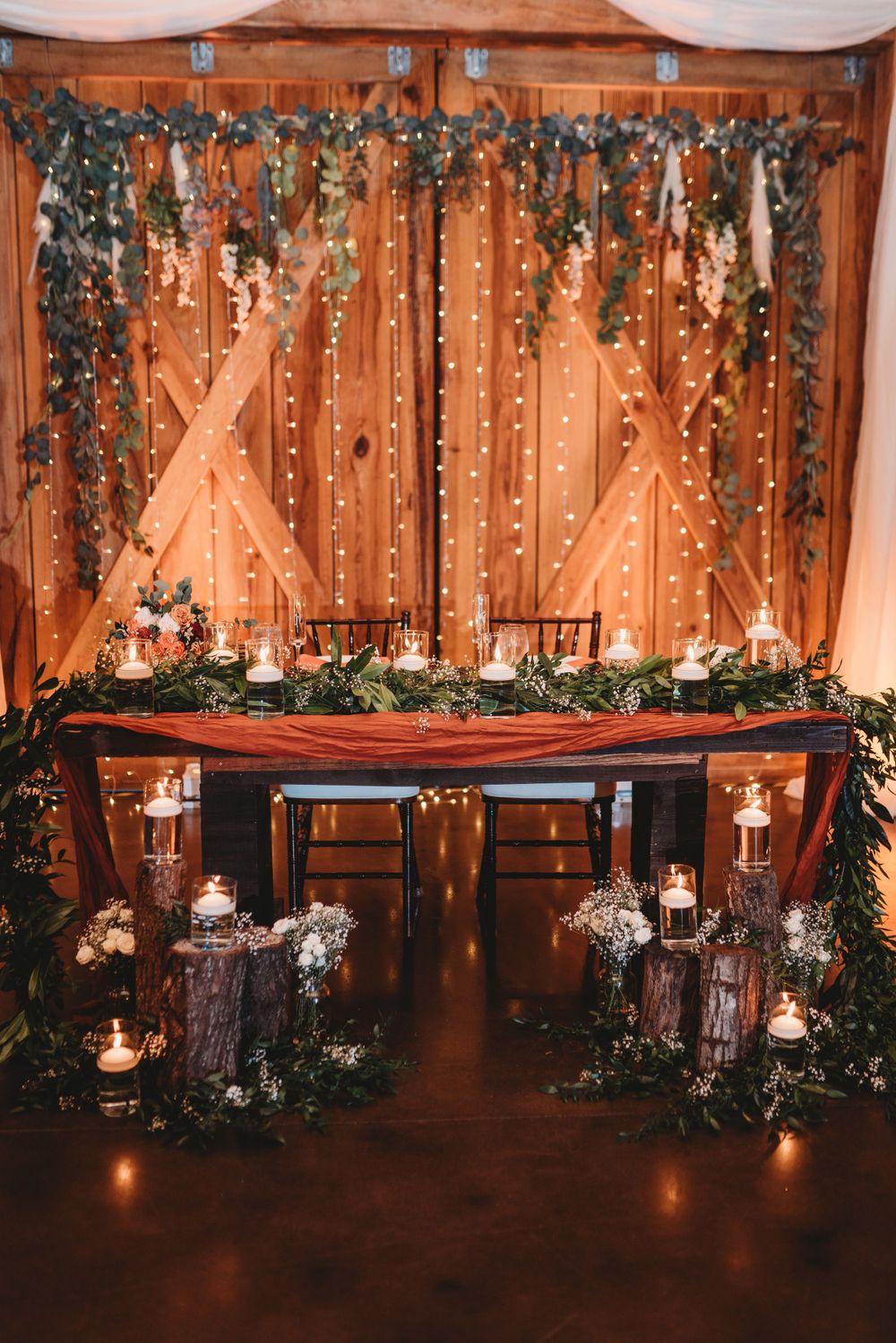 Rustic wedding head table decorated with fairy lights, greenery and candles against wooden barn doors.