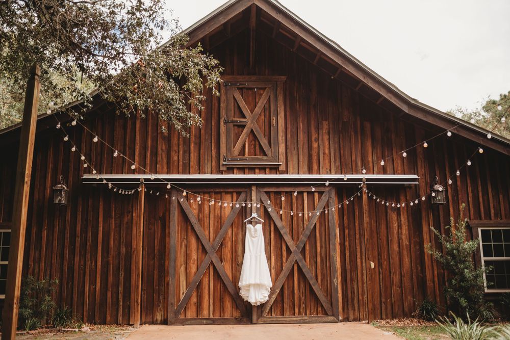 A classic white wedding dress hangs from a rustic wooden barn with string lights decorating the exterior.
