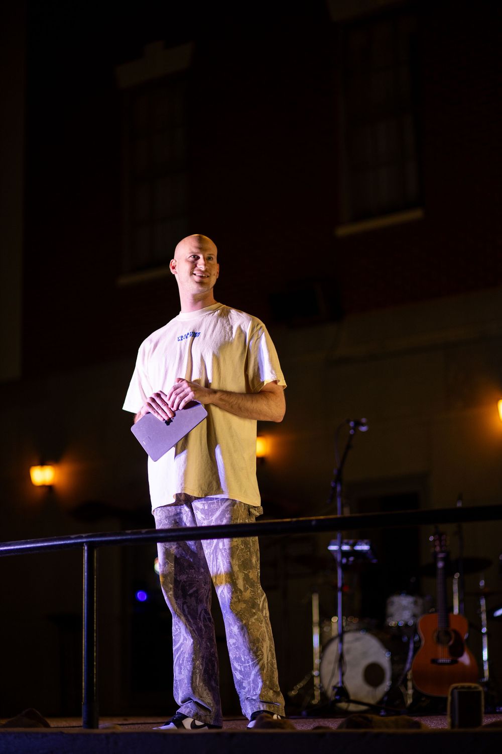 Bald man smiling on stage at night holding a notebook at an outdoor worship gathering.