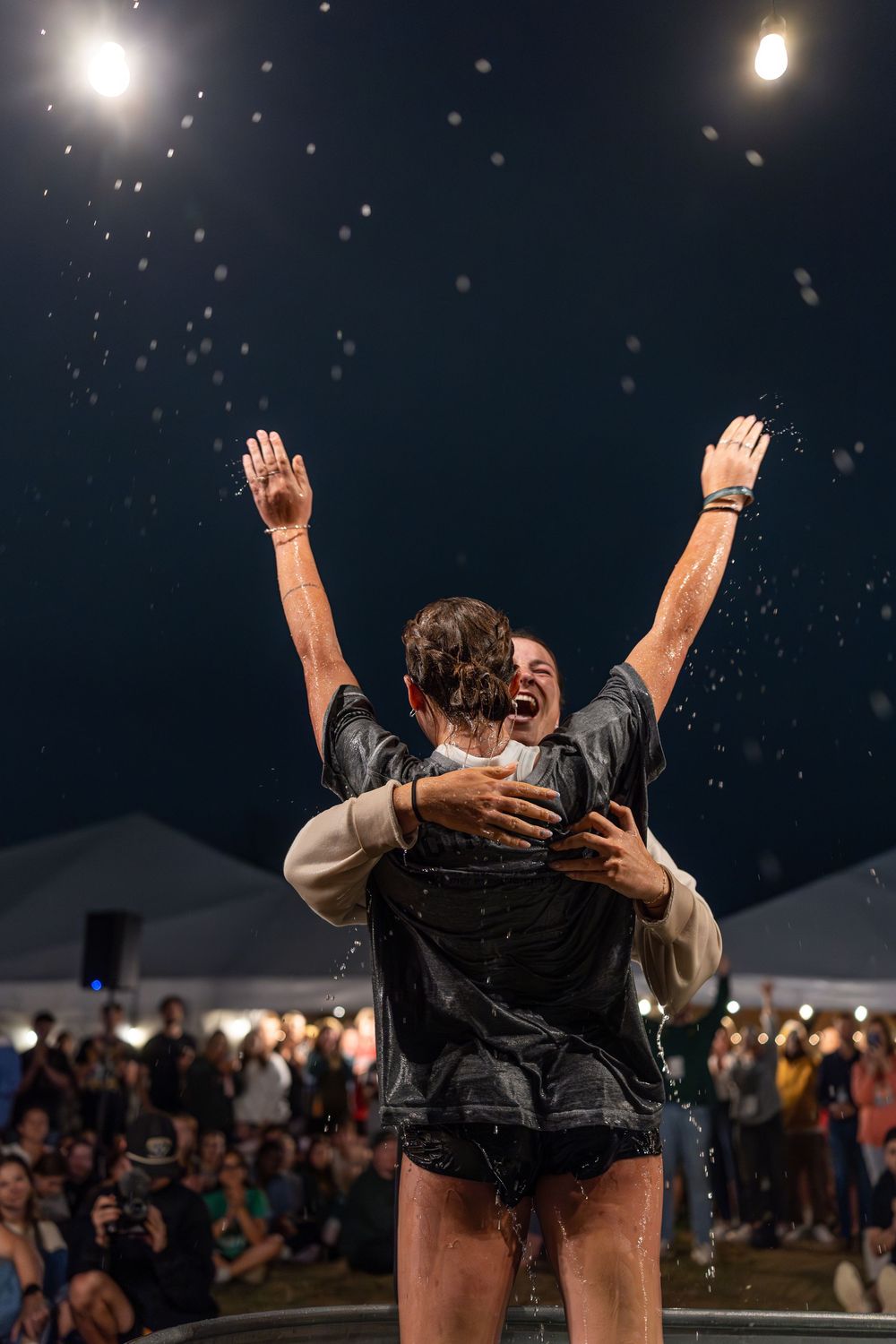 Joyful woman with arms raised being lifted after baptism outdoors at night event.