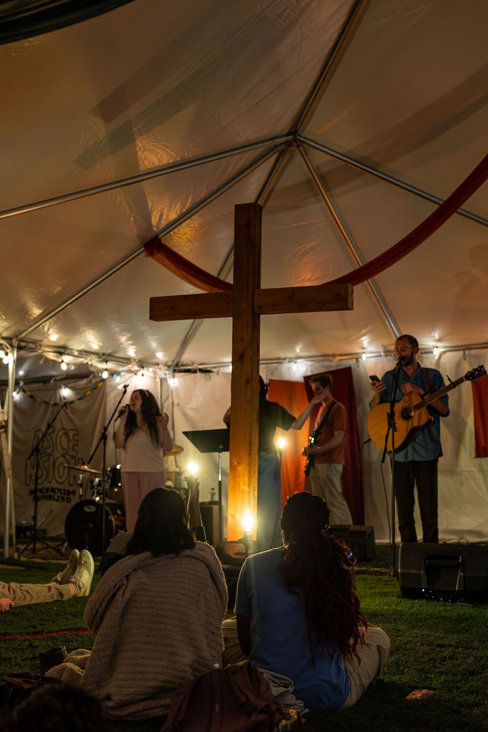 Small worship gathering inside tent with wooden cross and guitarist performing under warm lights.