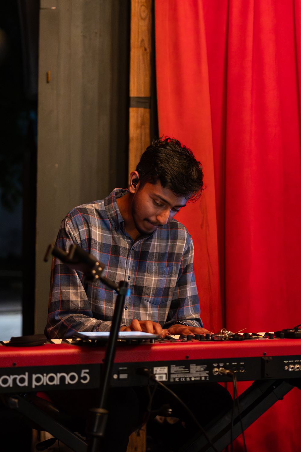 Young man playing a Nord Piano 5 keyboard on stage with a red curtain backdrop.