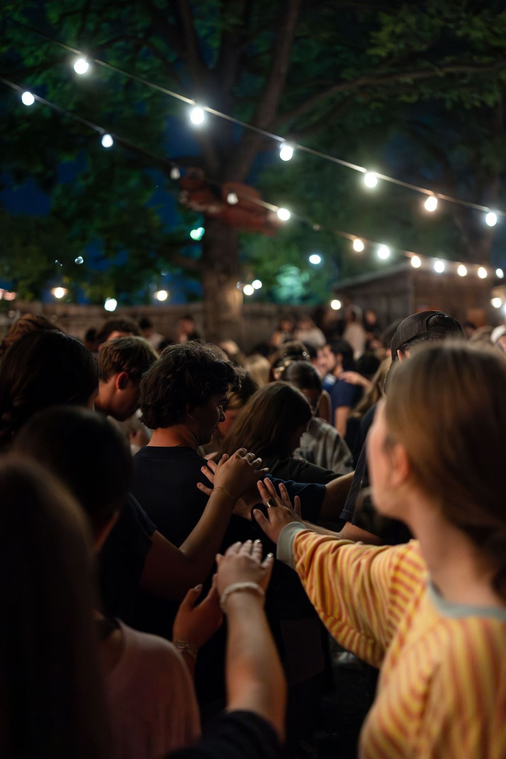 Crowd of young people praying together outdoors under string lights at a nighttime worship event.