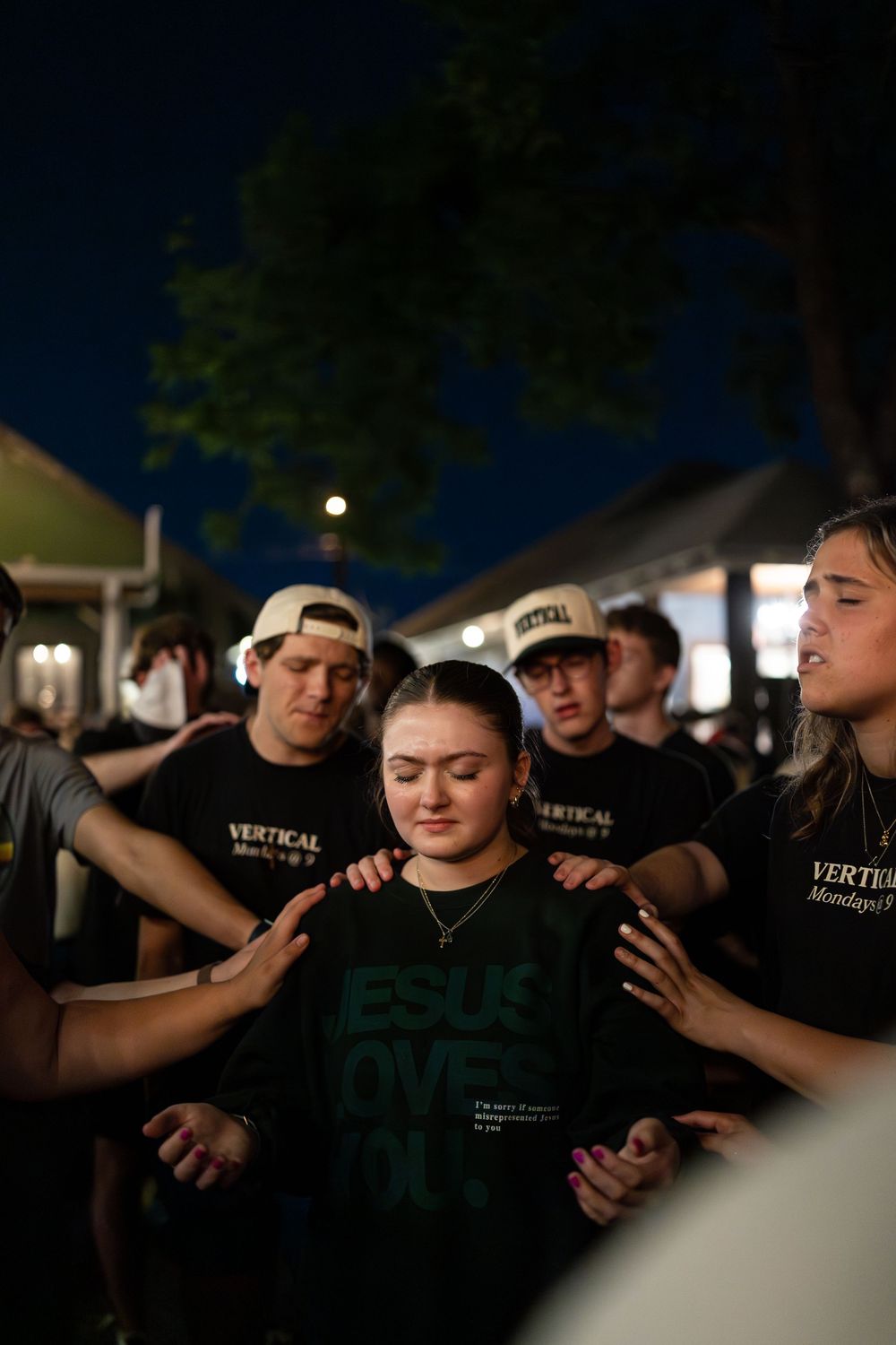 Group of young people in Vertical ministry shirts laying hands on a girl in prayer outdoors.