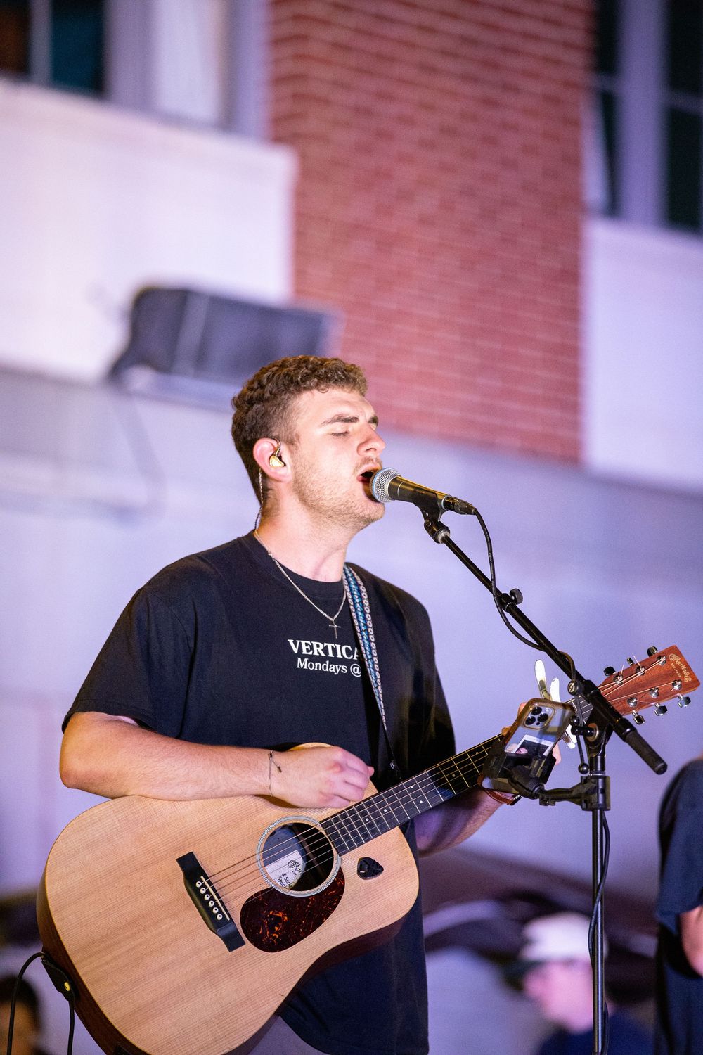 Young man in a Vertical ministry shirt singing and playing acoustic guitar at an outdoor event.