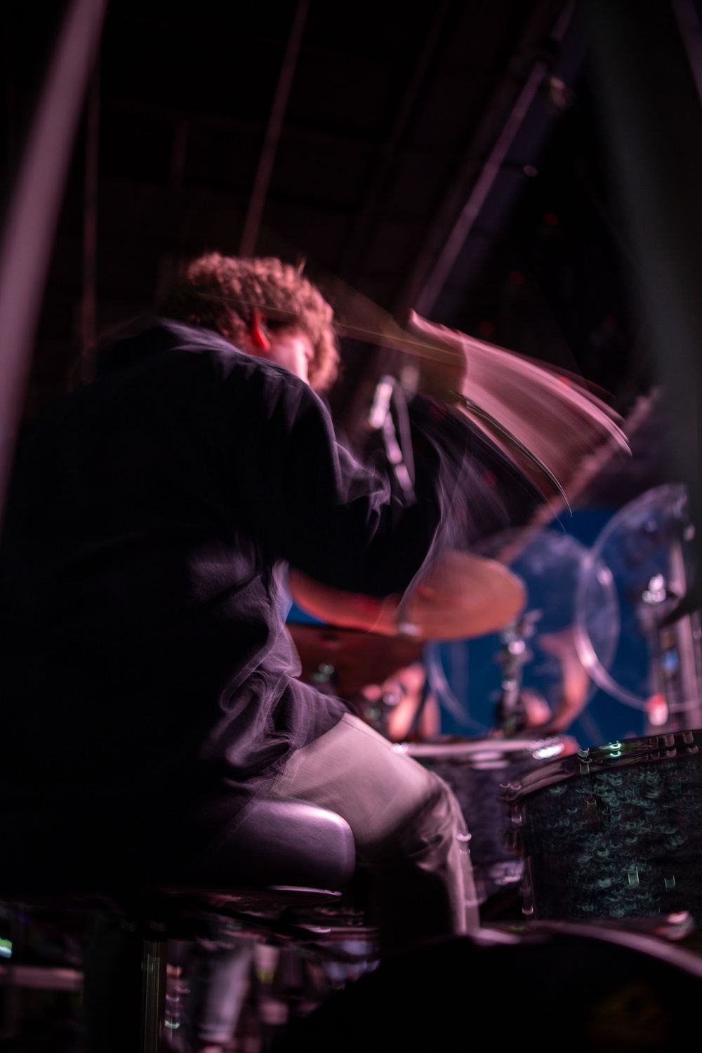 Dynamic motion blur shot of drummer performing on stage under purple and red concert lighting.