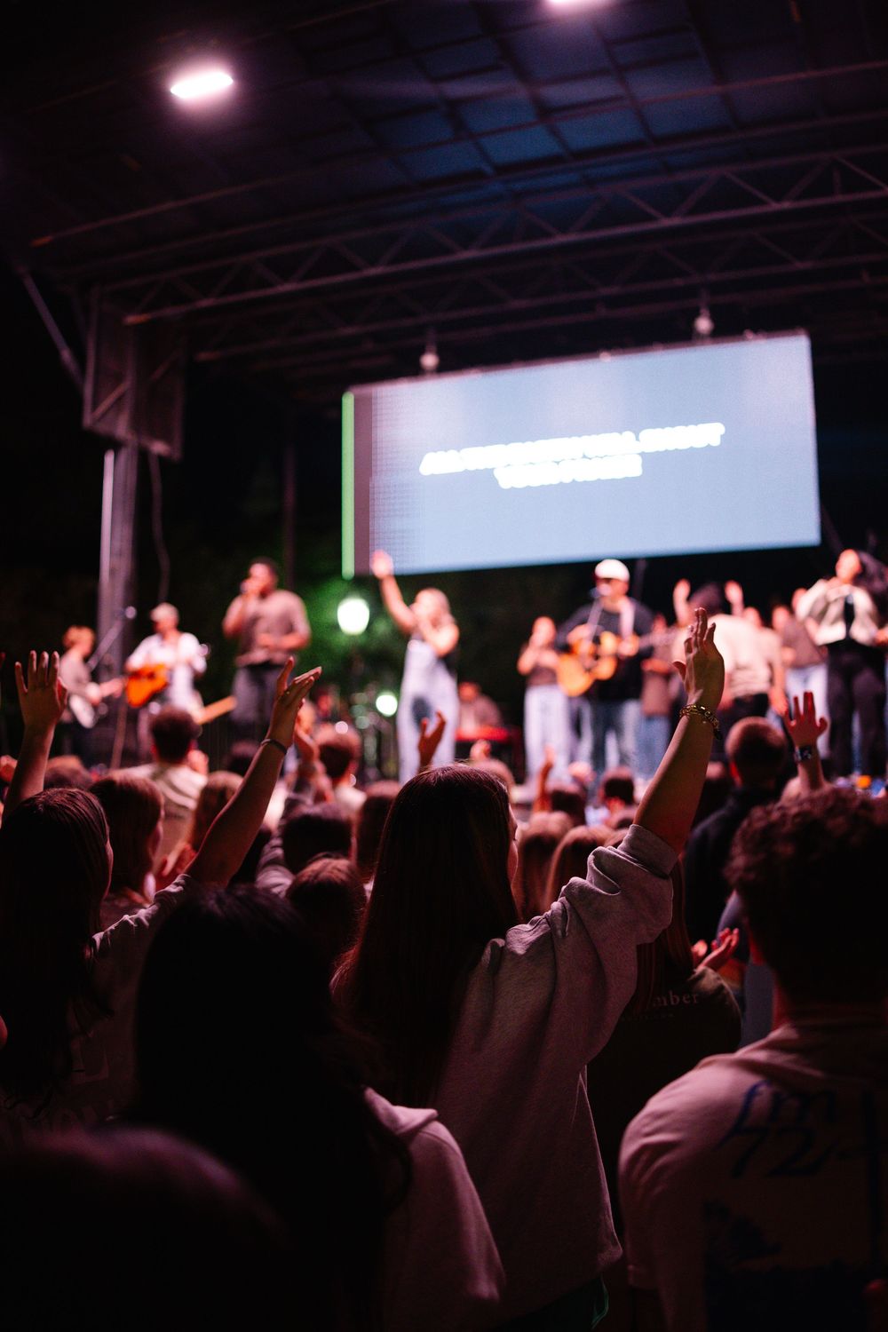 Crowd with raised hands worshipping at an outdoor Christian concert with performers on lit stage.