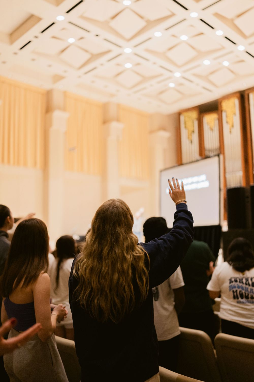 Students raise hands in worship during a college chapel service in an elegant auditorium.