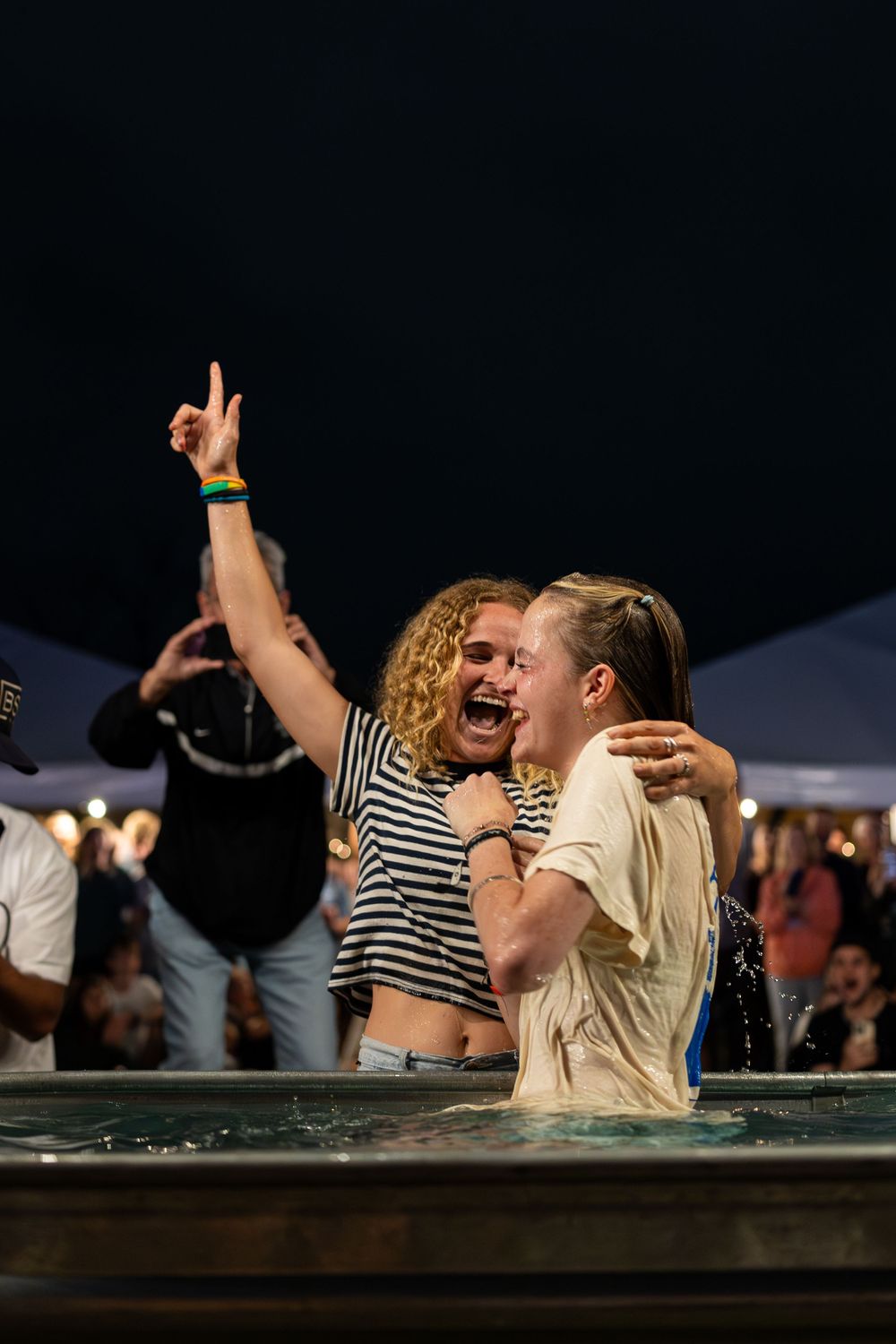 Two young women celebrating joyfully together in baptismal pool at outdoor nighttime Christian event.