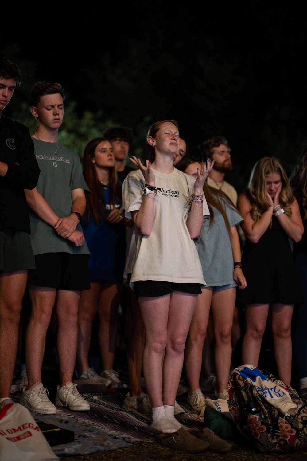 Young woman with hands raised in worship surrounded by a group of students at a nighttime event.