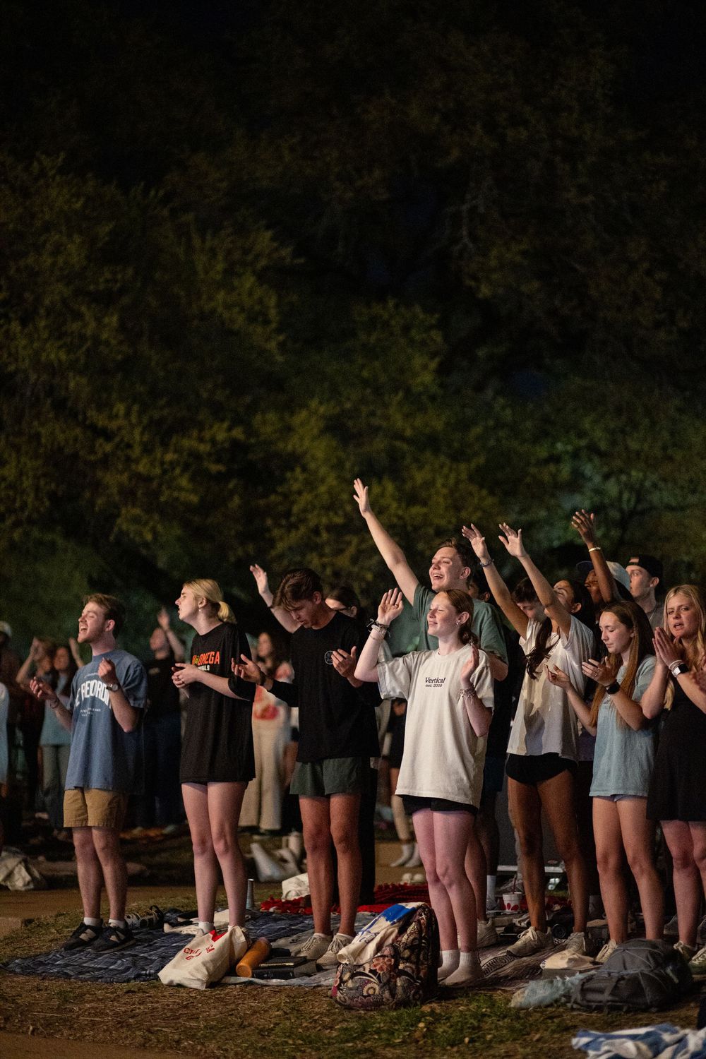 Group of young people raising hands in worship at an outdoor nighttime Christian gathering.