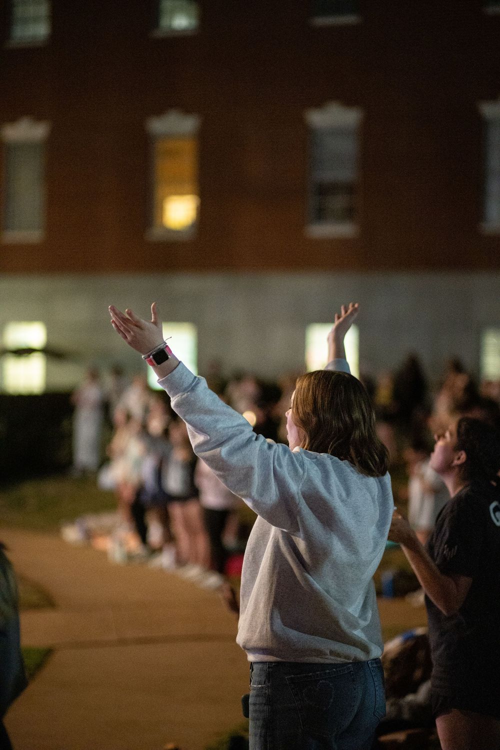 Young woman in a gray sweatshirt raising both hands in worship at an outdoor nighttime event.