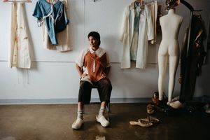 Model sitting in a chair against a fashion collection hung on the wall, and a mannequin