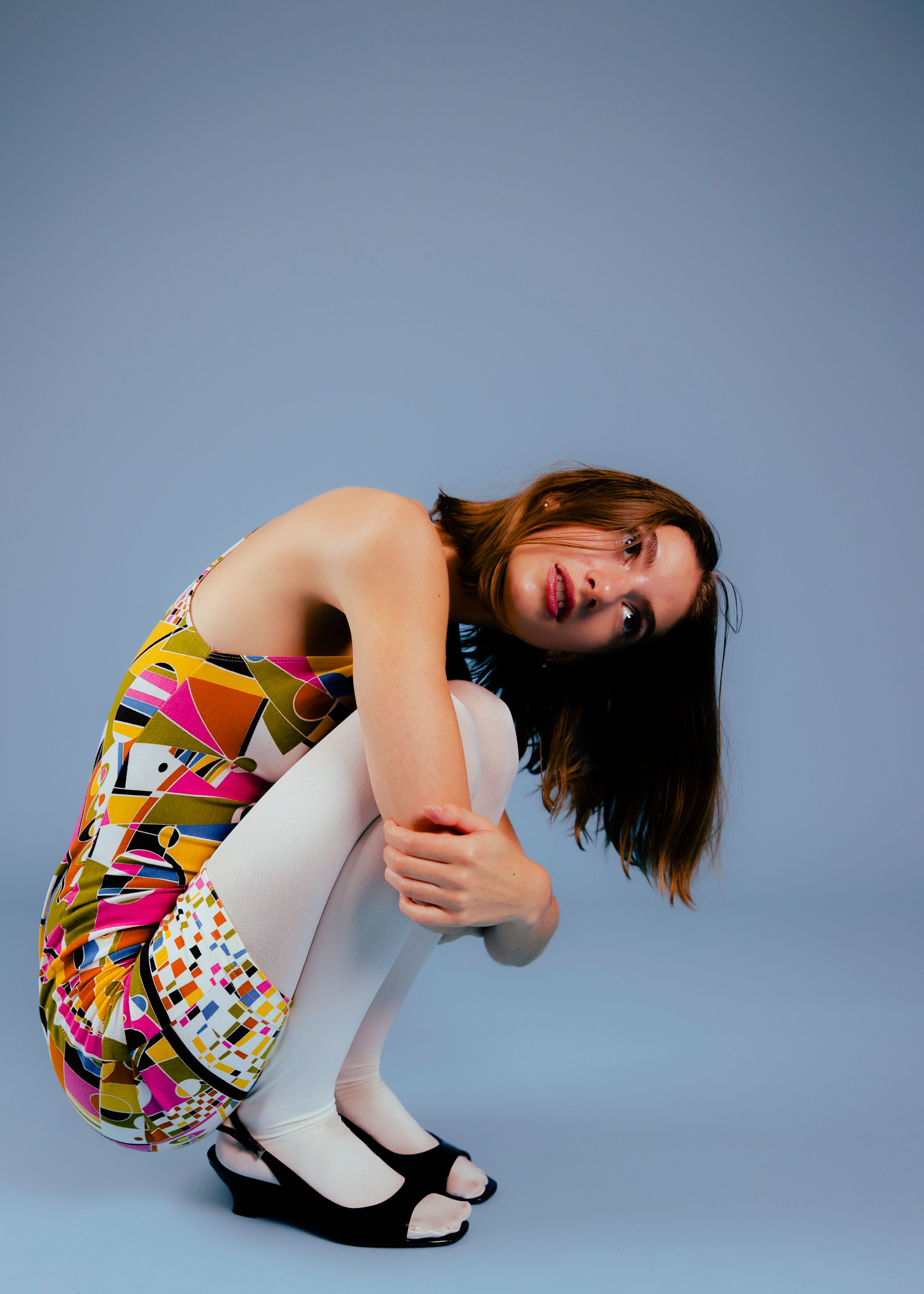 Model posed in 70s style dress against a blue backdrop, holding her knees