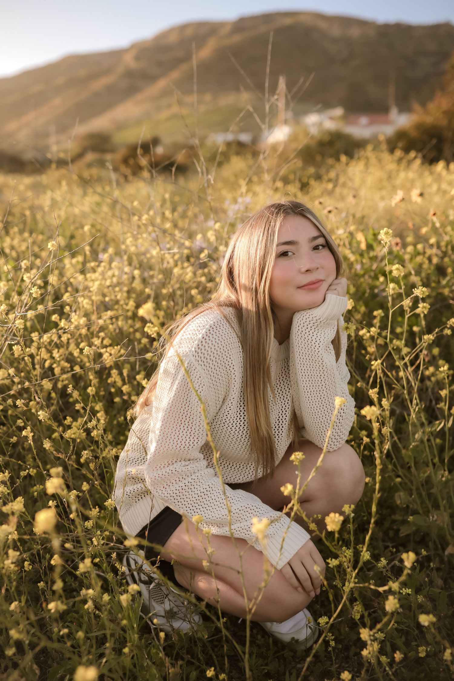 Someone wearing a cream sweater crouches in a field of yellow wildflowers at golden hour in the countryside.