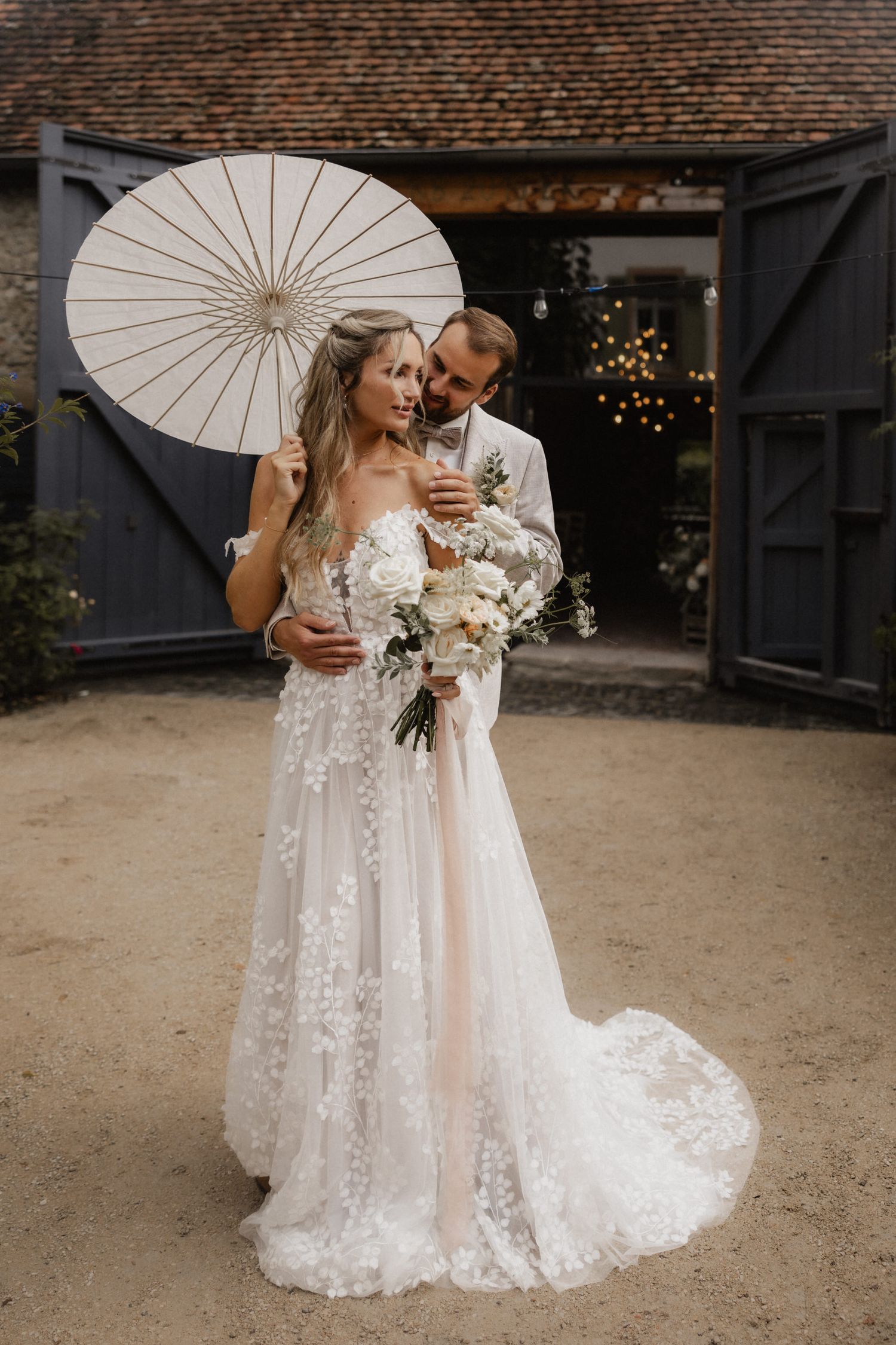 Couple poses with white parasol and floral bouquet against rustic backdrop.