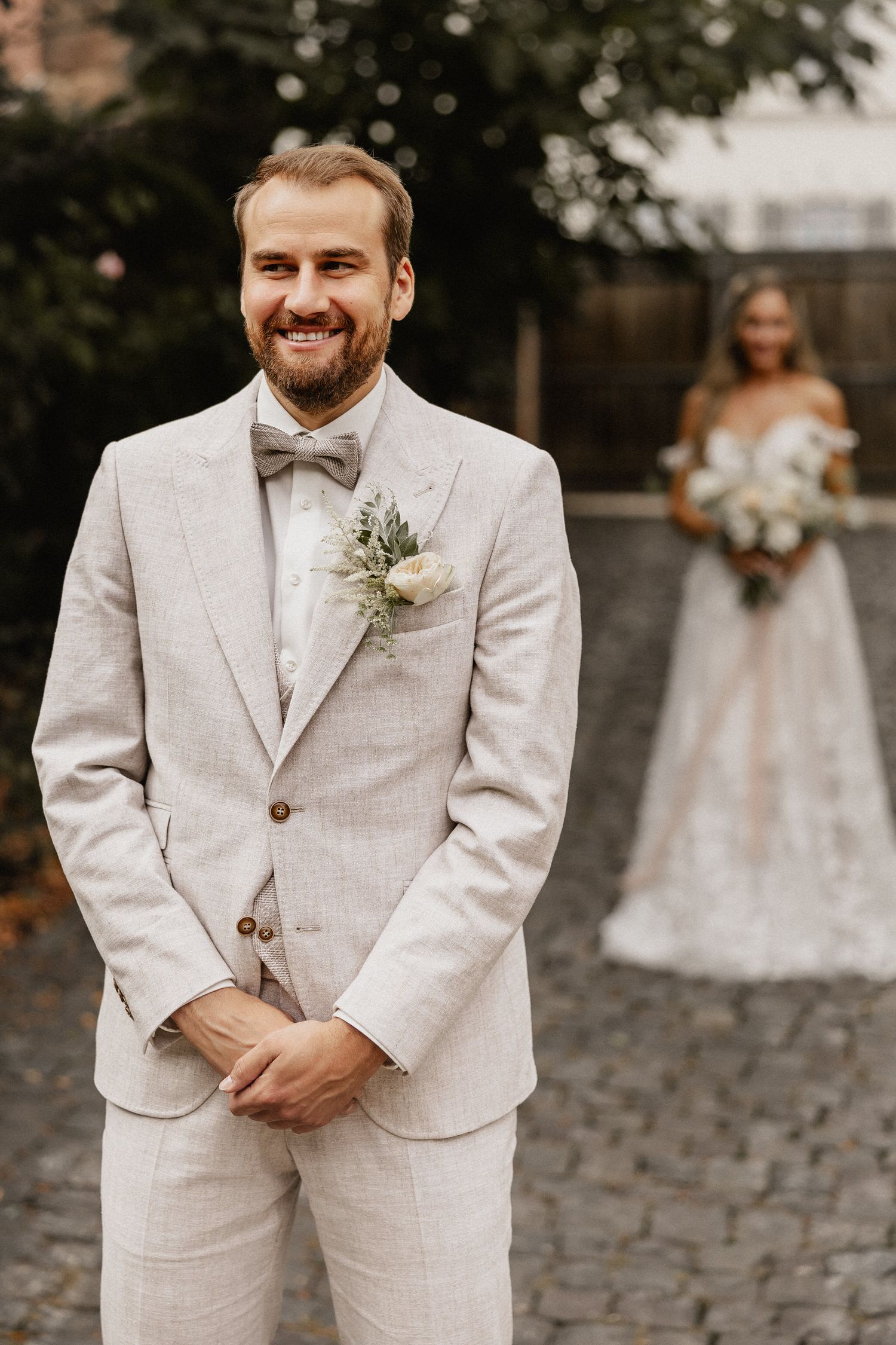 Groom in light beige suit and bow tie standing on cobblestone path during outdoor wedding.