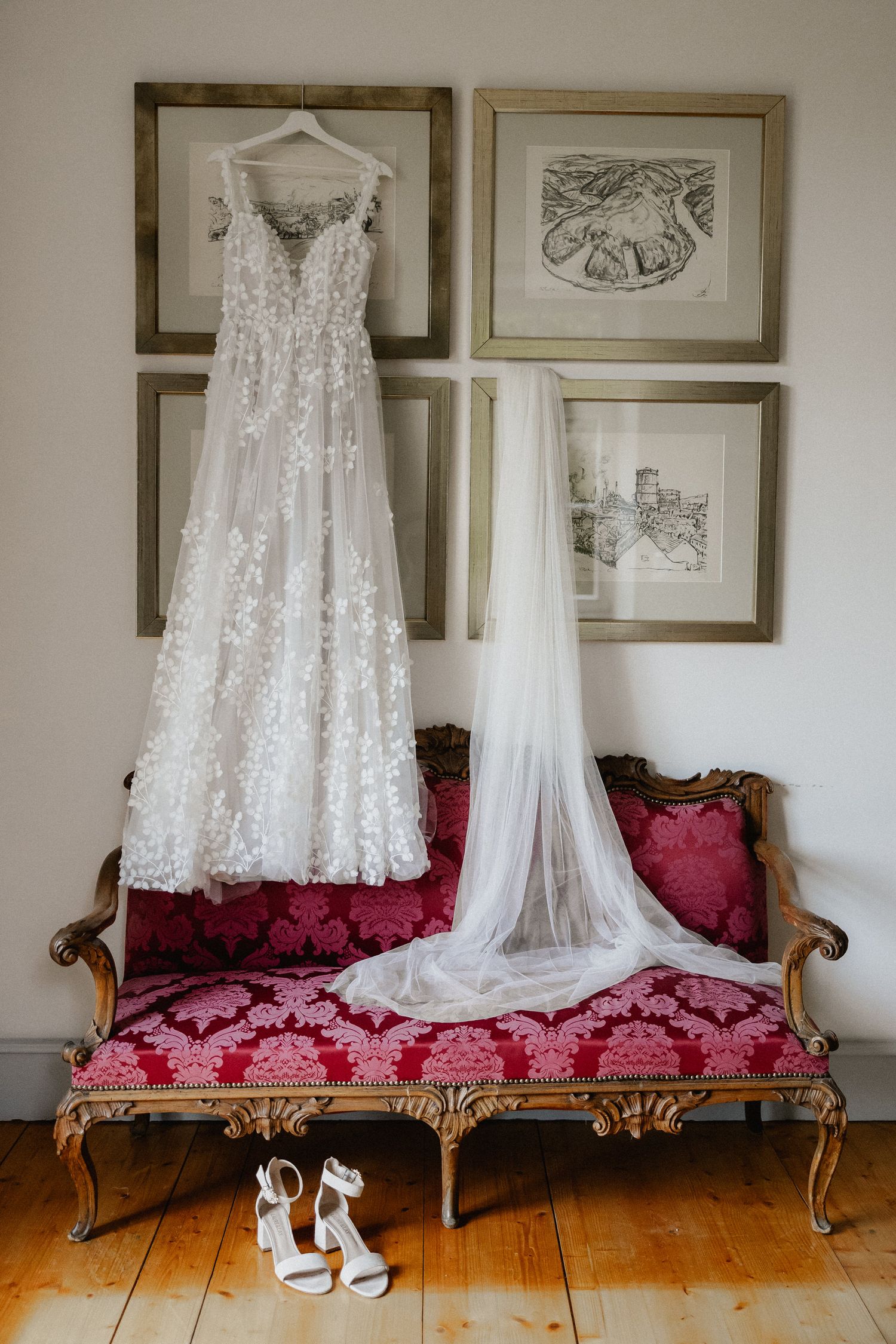 White wedding dress and shoes displayed on vintage red velvet settee against wall with framed artwork.