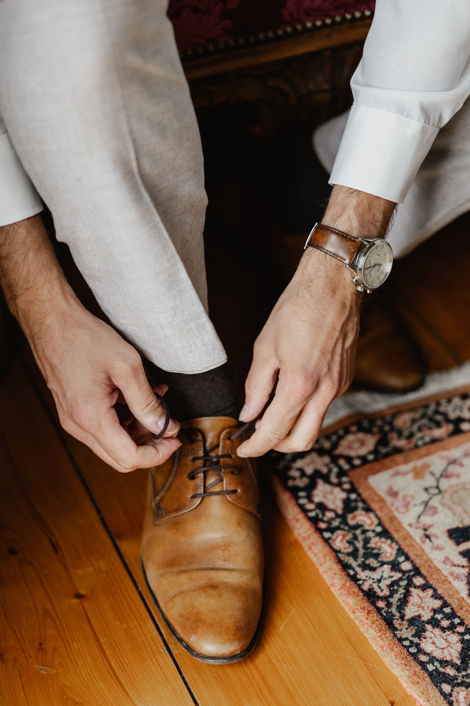 Hands tying brown leather dress shoes on wooden floor next to oriental rug.