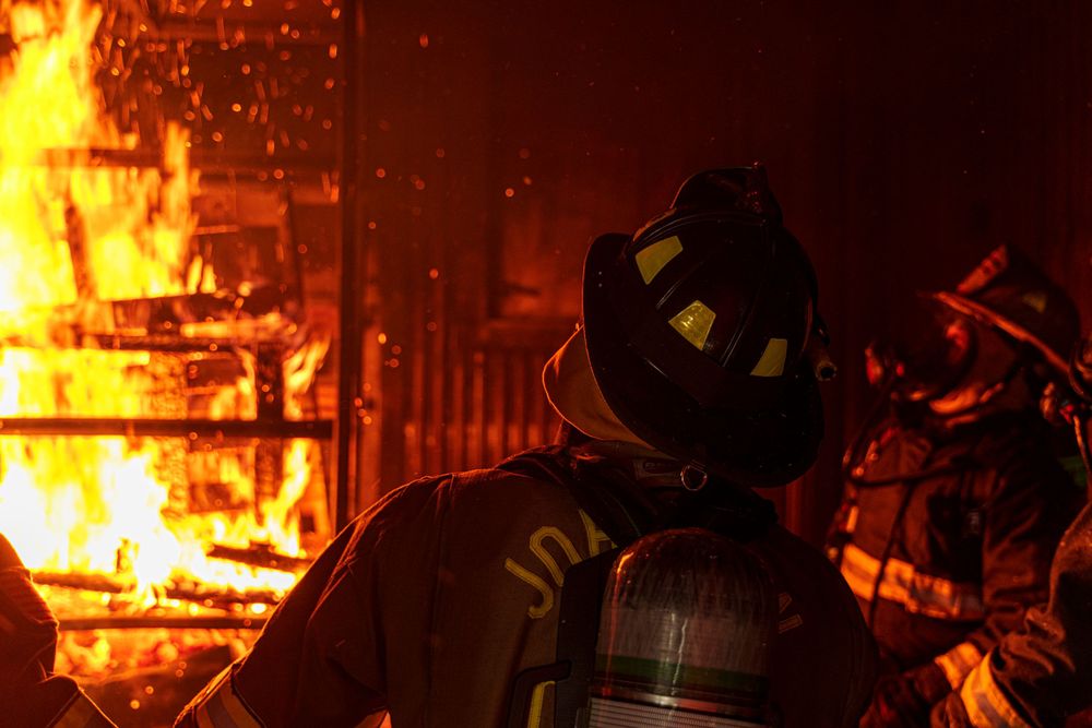 Firefighter observes raging flames through protective gear during nighttime emergency.