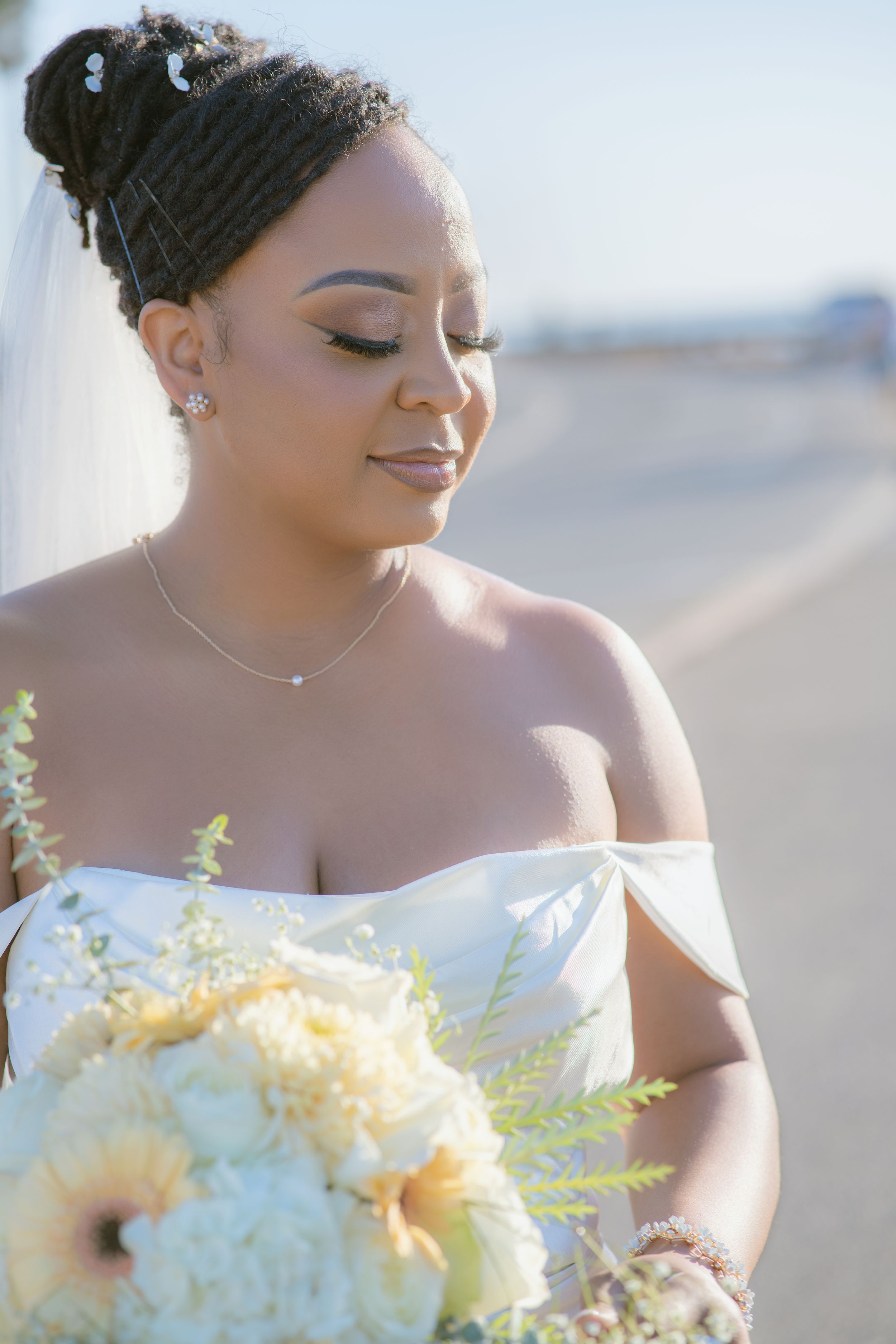 A bride in an elegant off-shoulder white gown holds a bouquet during a sunset photoshoot.
