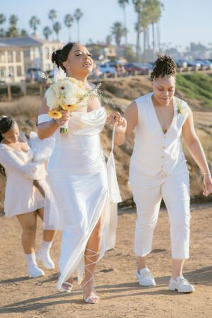 Wedding party wearing all white walks along a coastal path with palm trees in the background.