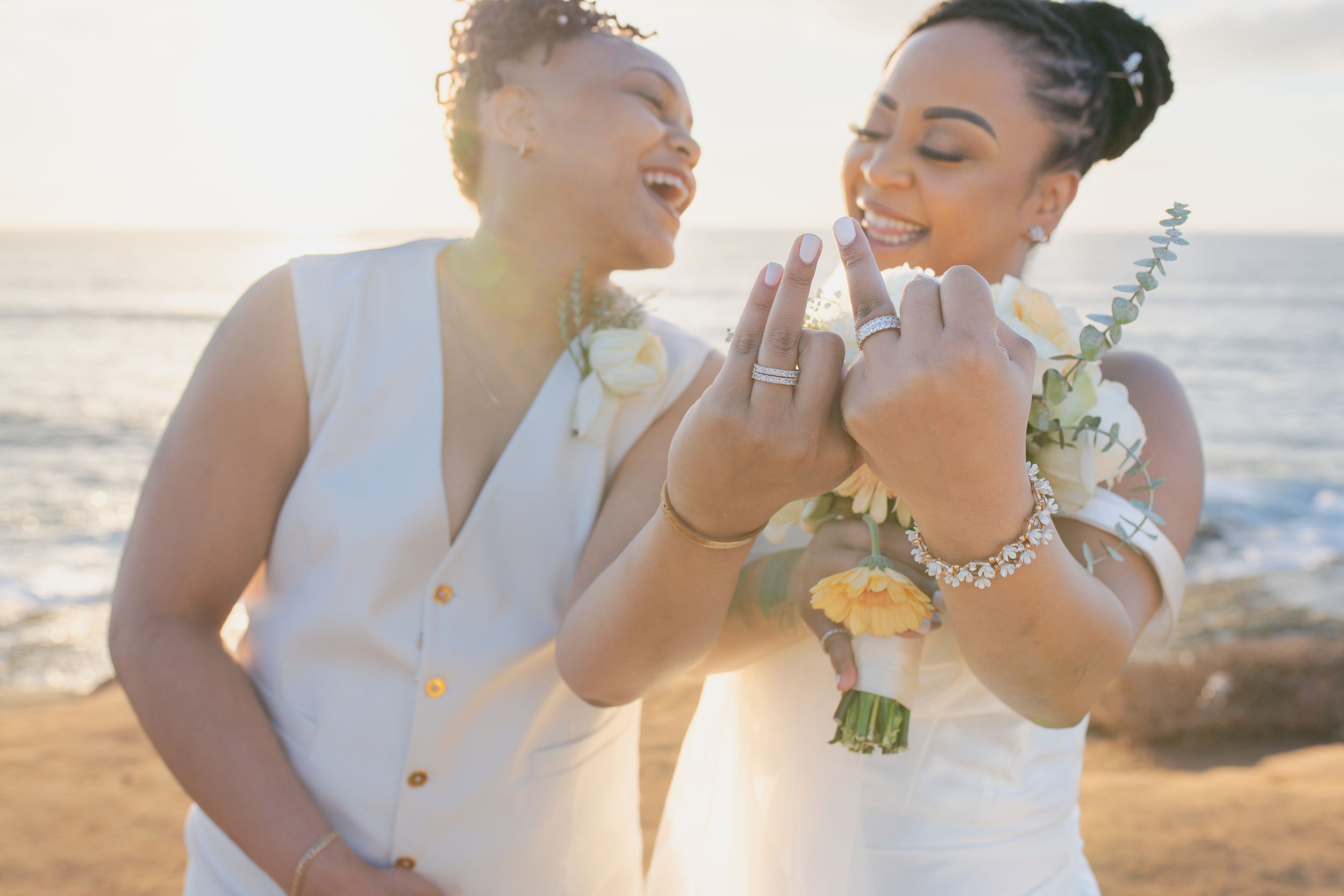 Newlyweds in white outfits share a joyful moment while showing off their rings at the beach.