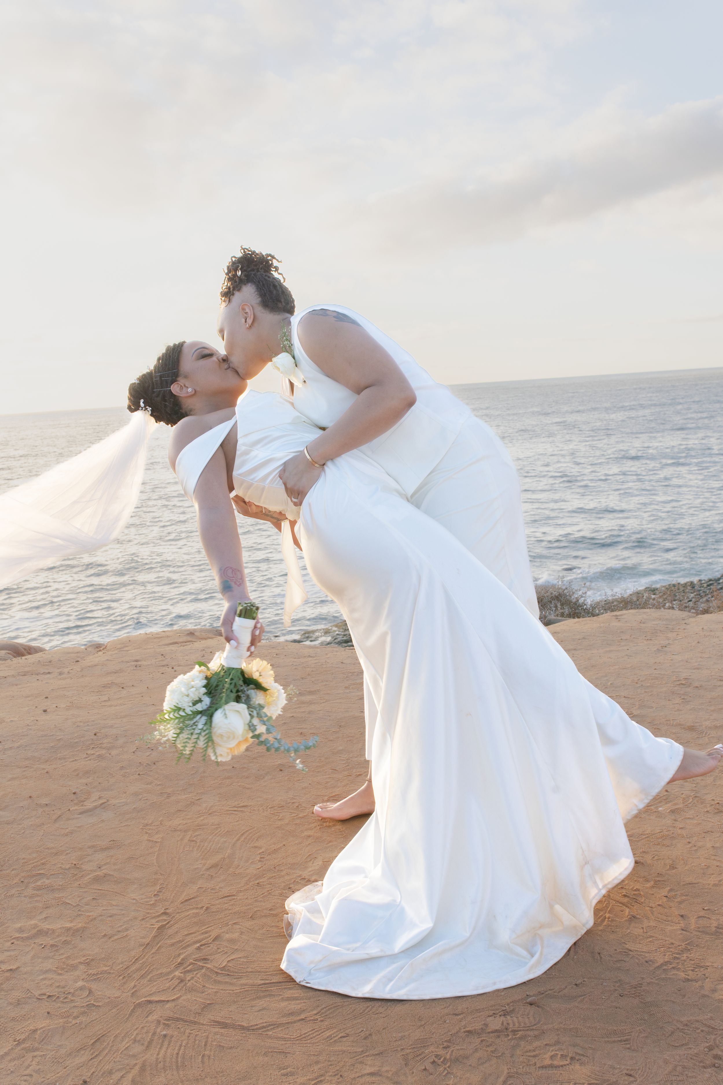 A romantic dip pose on the beach with white wedding attire and bouquet at sunset.