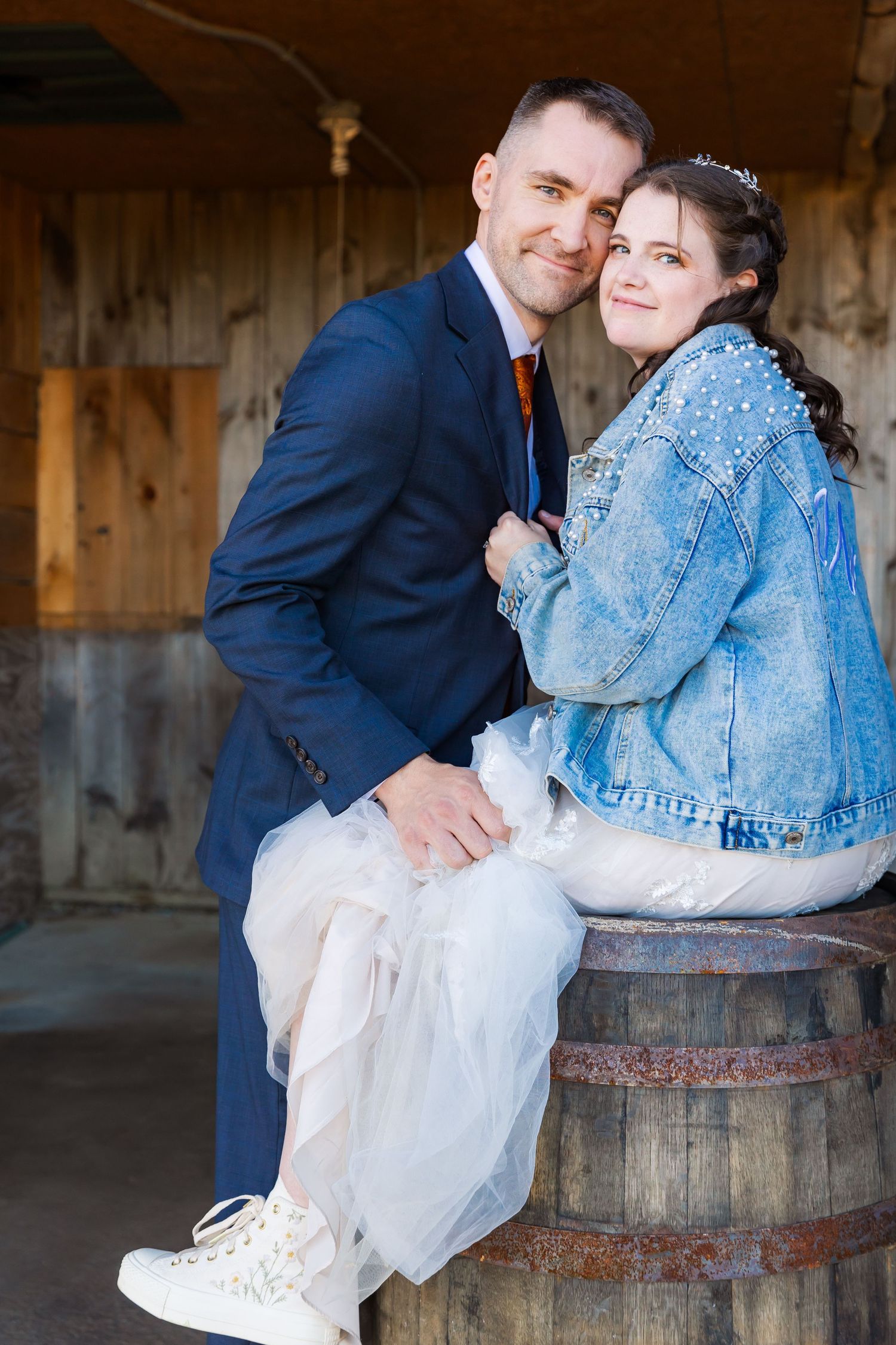 Couple in wedding attire posing together on rustic wooden barrel in barn setting.