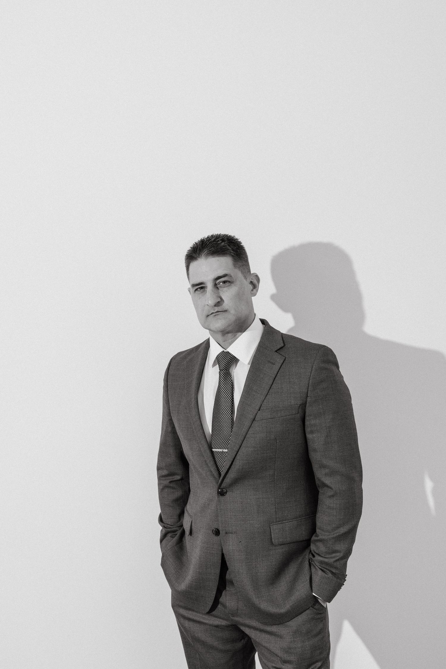 Black and white portrait of a professional man in a suit and tie standing confidently against a white wall.