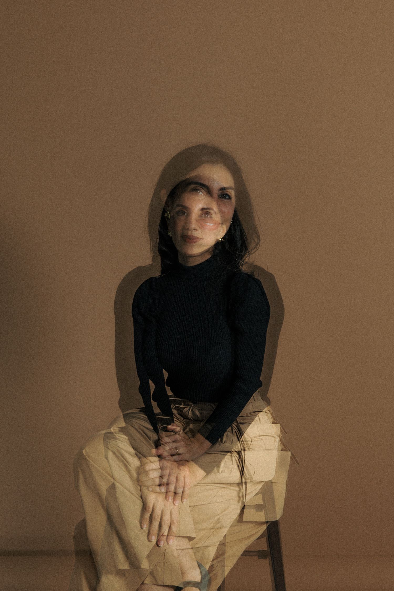 A person in a black turtleneck and beige pants sits on a chair against a brown backdrop in moody lighting.