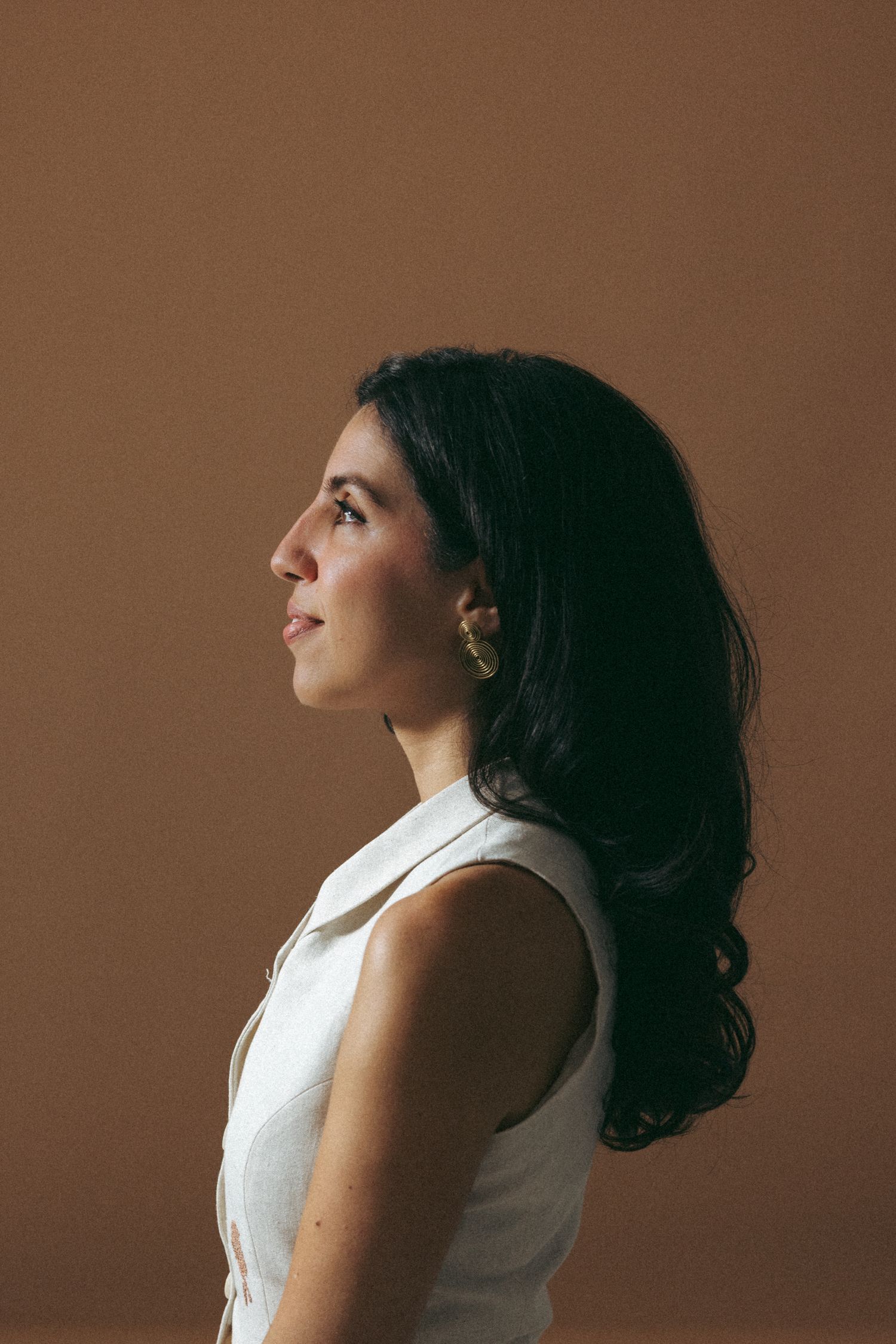 Woman with dark curly hair in white sleeveless top poses against brown backdrop in artistic profile portrait series.