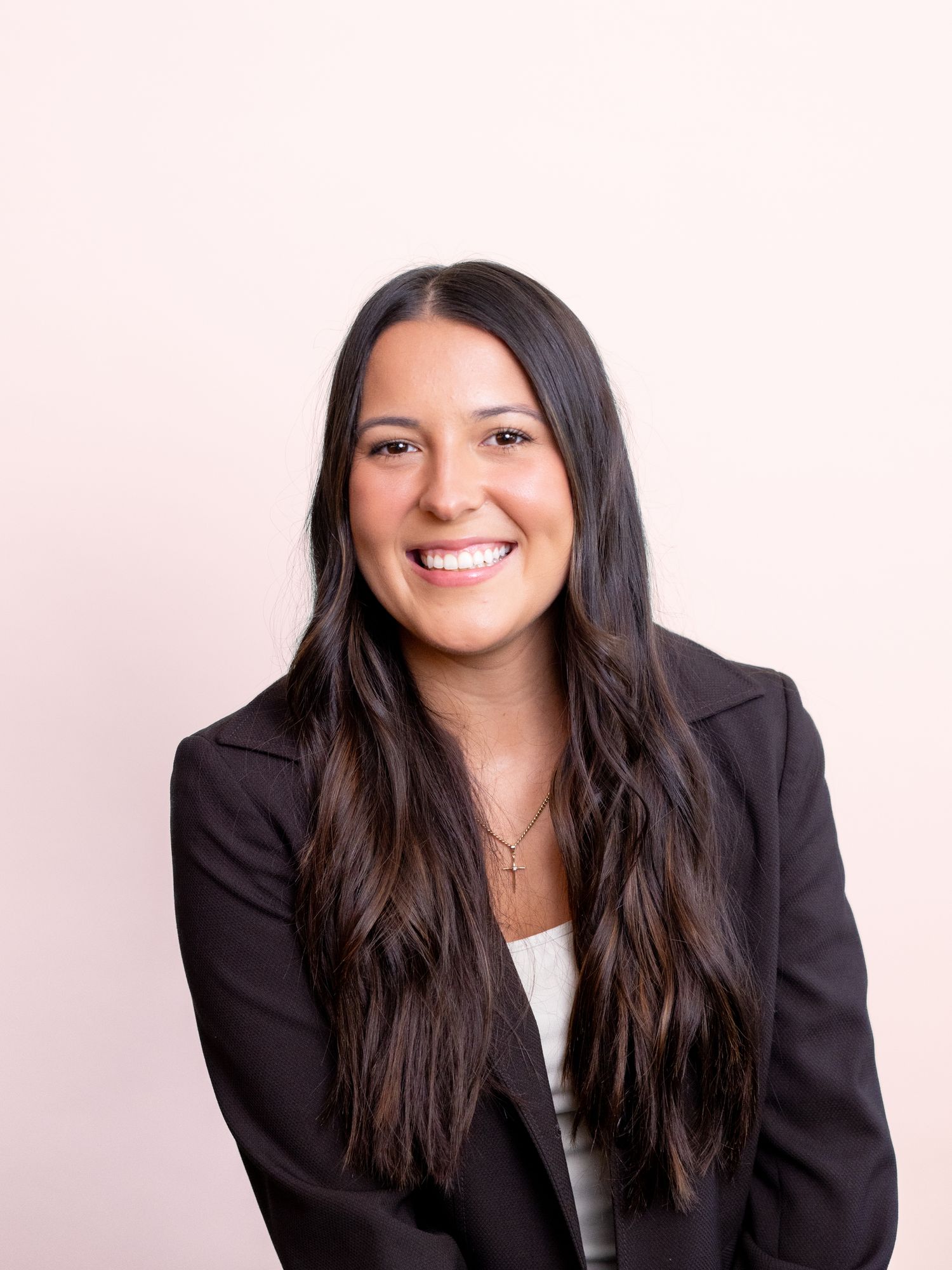 Professional woman with long dark hair smiling in a black blazer against a light pink background.