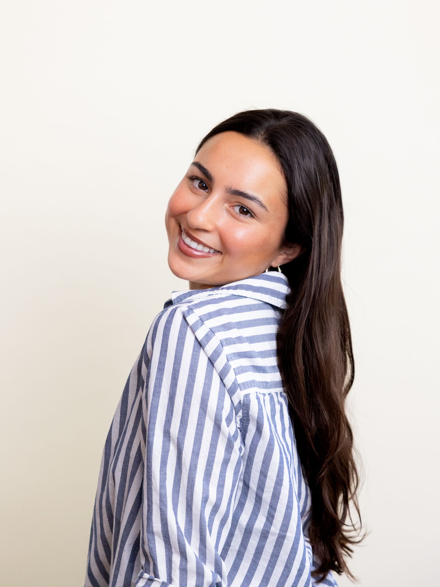 Smiling young woman with long dark hair wearing a blue and white striped shirt, posing against a light background.