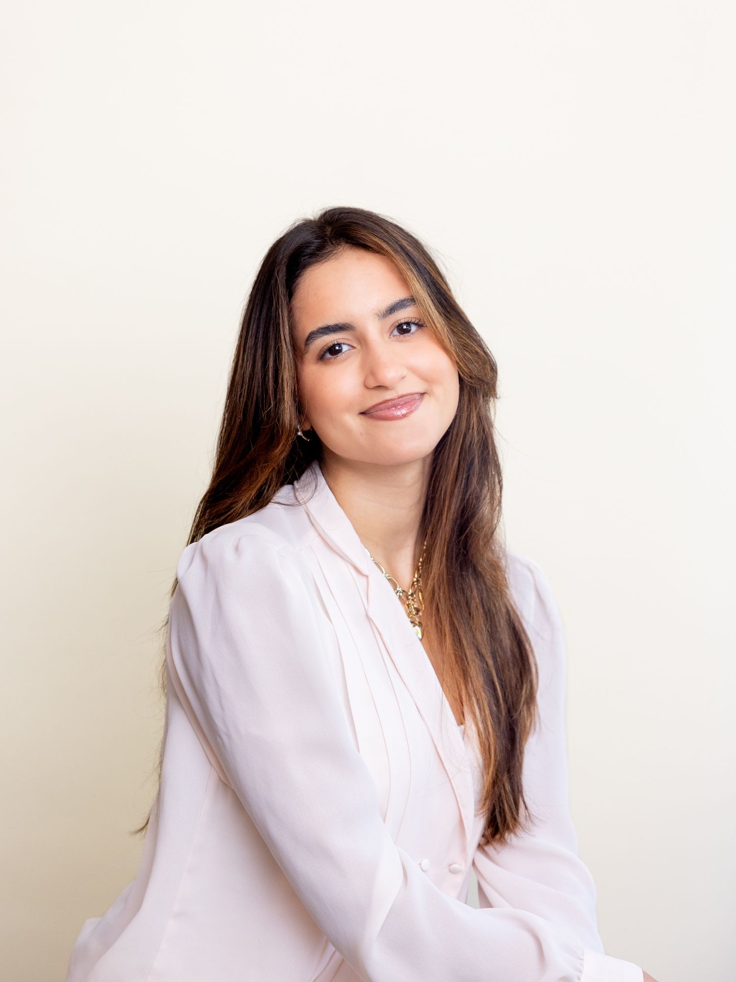 Young woman with long brown hair wearing a white blouse and gold necklace, smiling against a cream background.