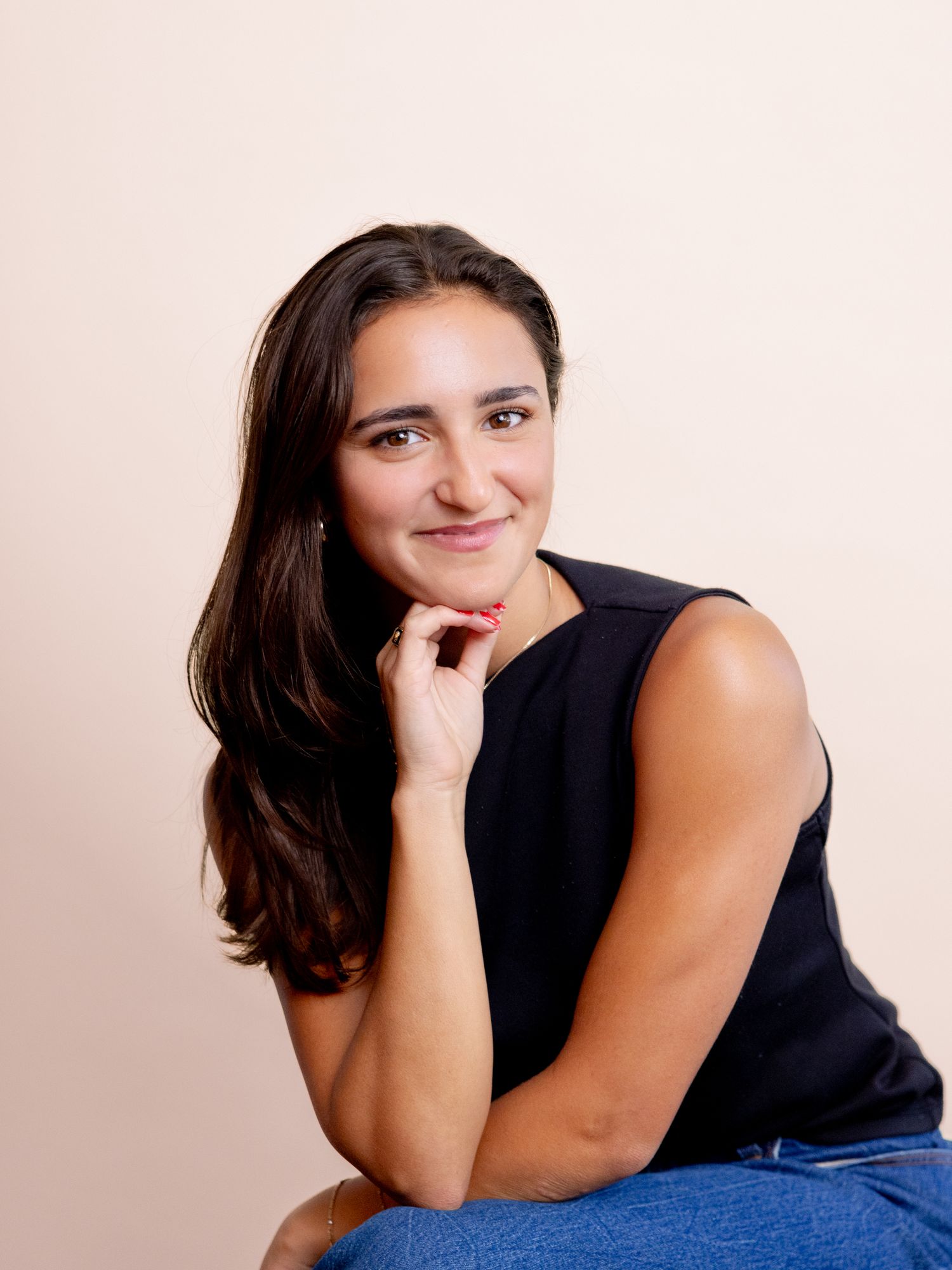 Young woman with dark hair in black sleeveless top and jeans, sitting with hand on chin, smiling softly.