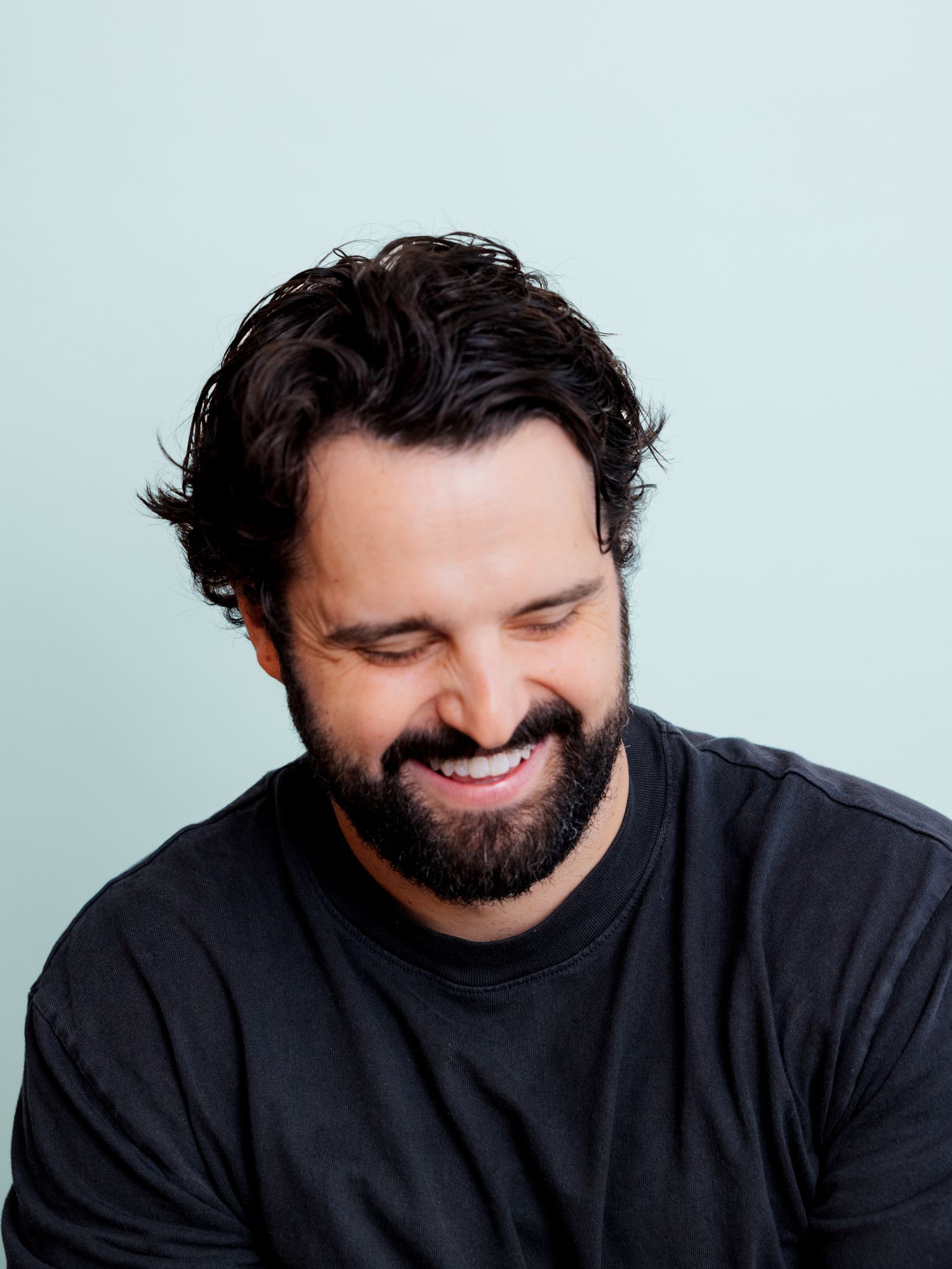 Smiling bearded man with dark curly hair wearing a black t-shirt against a light blue background.