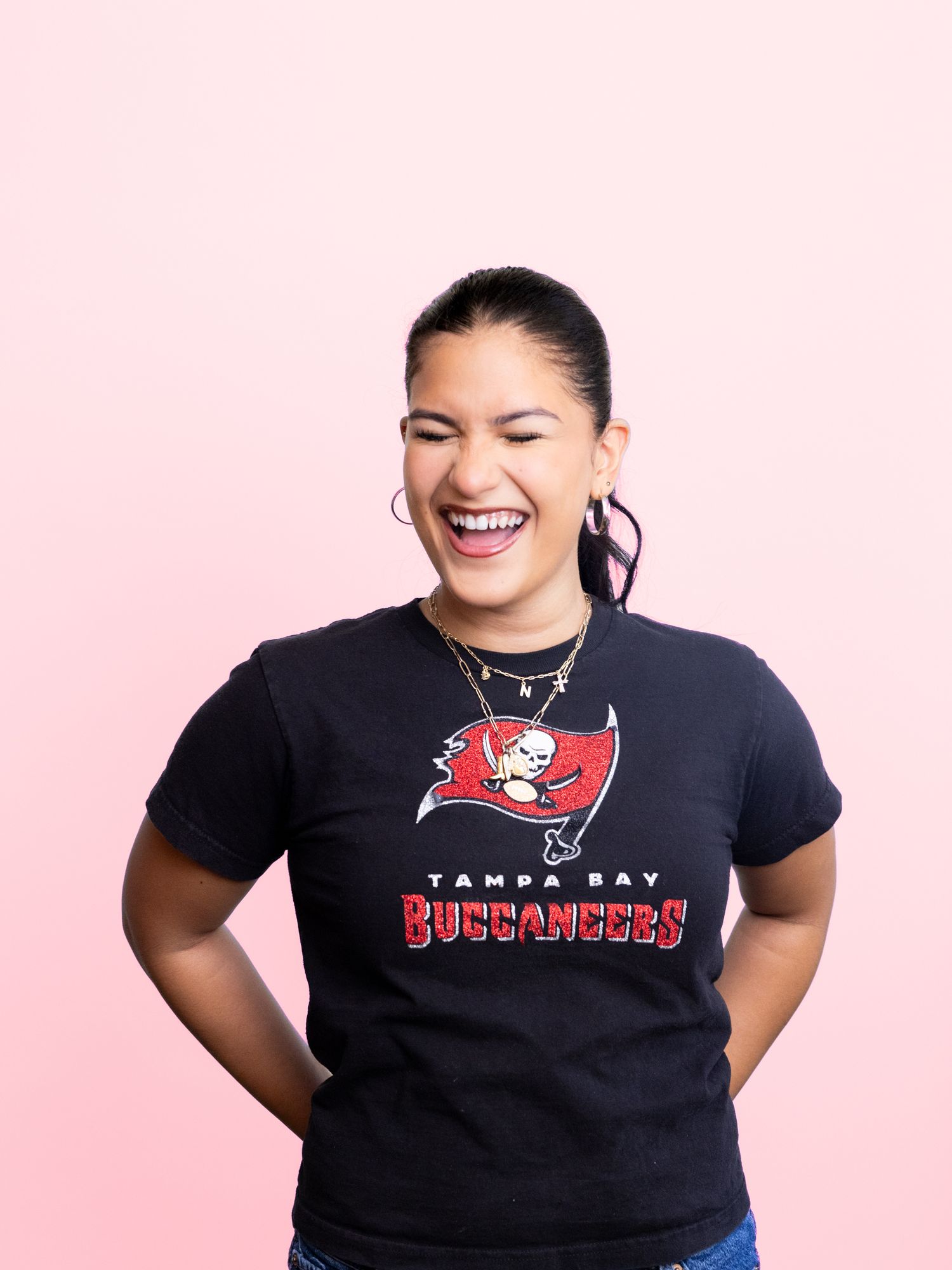 Smiling woman wearing a black Tampa Bay Buccaneers NFL t-shirt, posing against a pink background.