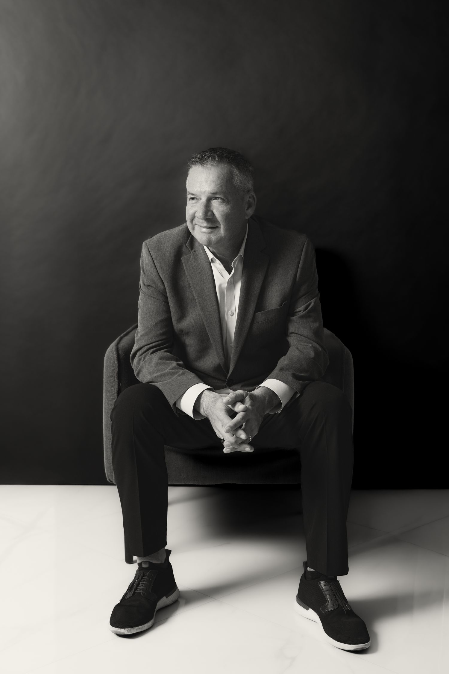 Black and white portrait of a smiling middle-aged man in a suit sitting relaxed in a chair against dark background.