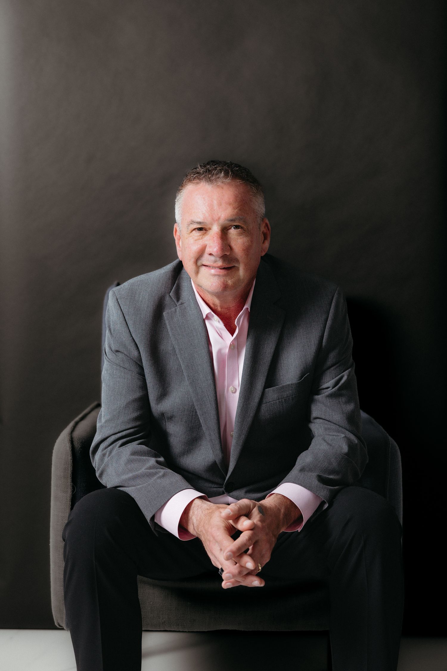 Professional businessman in gray suit and pink shirt seated confidently in chair against dark background.
