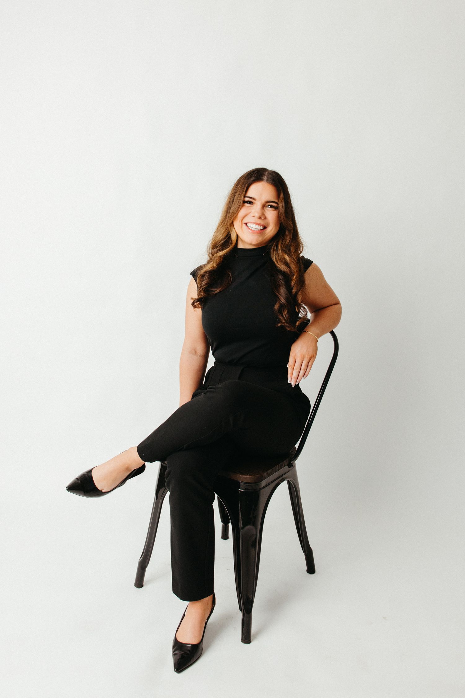 Professional woman in black outfit sits elegantly on metal chair against white background.